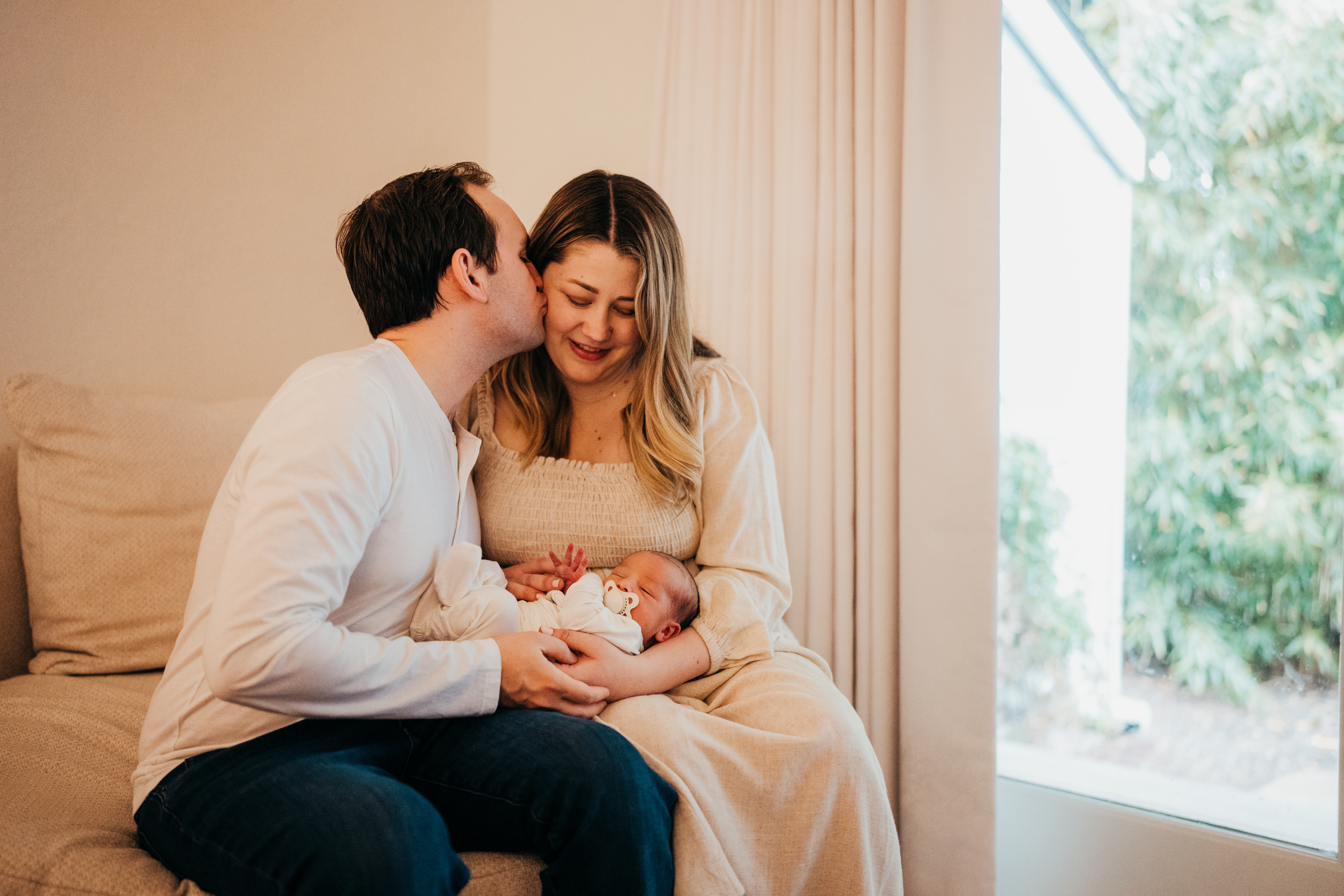 New dad kisses the cheek of his wife as they cradle their newborn on a couch in a window after using baby shower venues in seattle