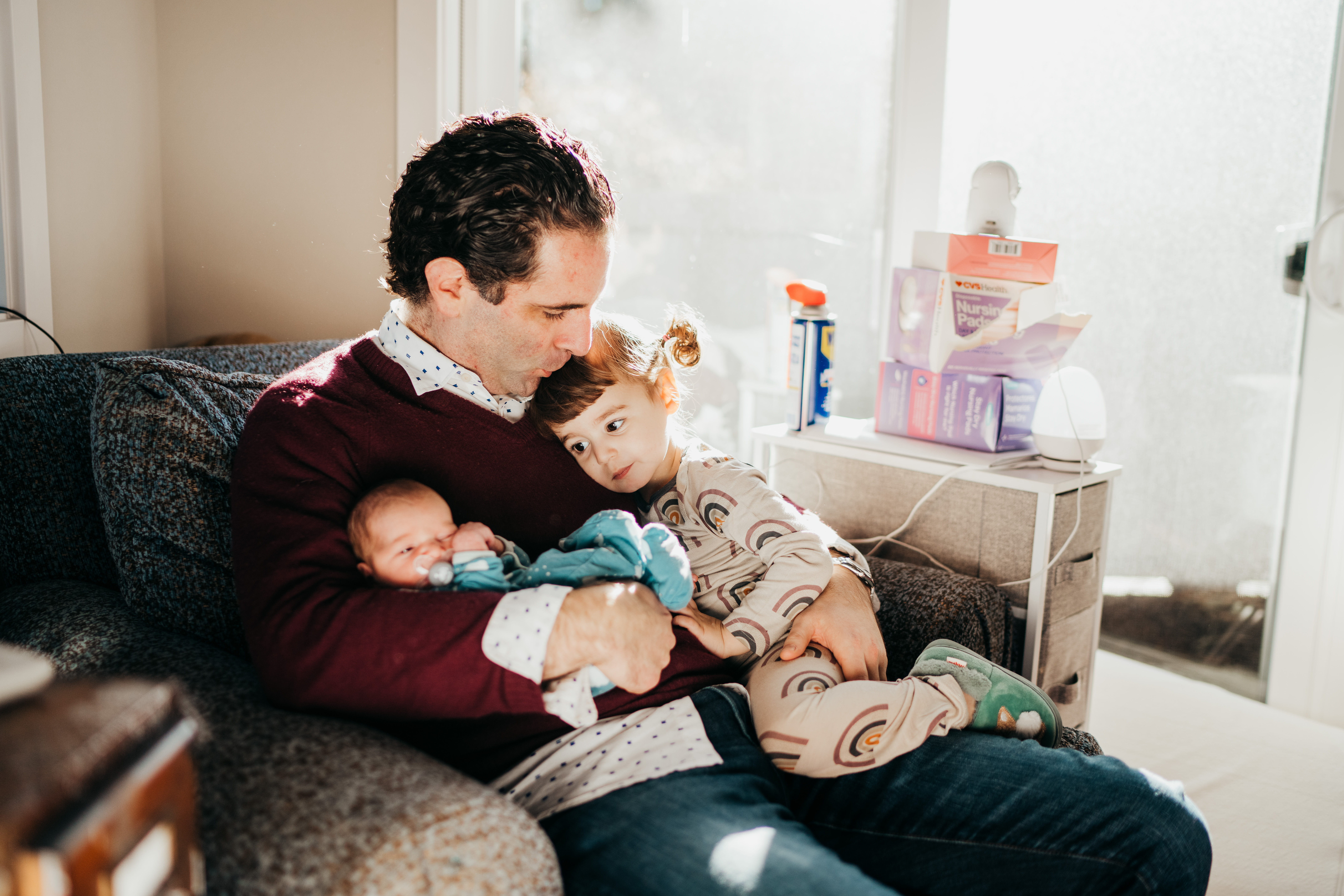 A dad kisses his toddler daughter's head while sitting in a nursing chair with newborn in other arm