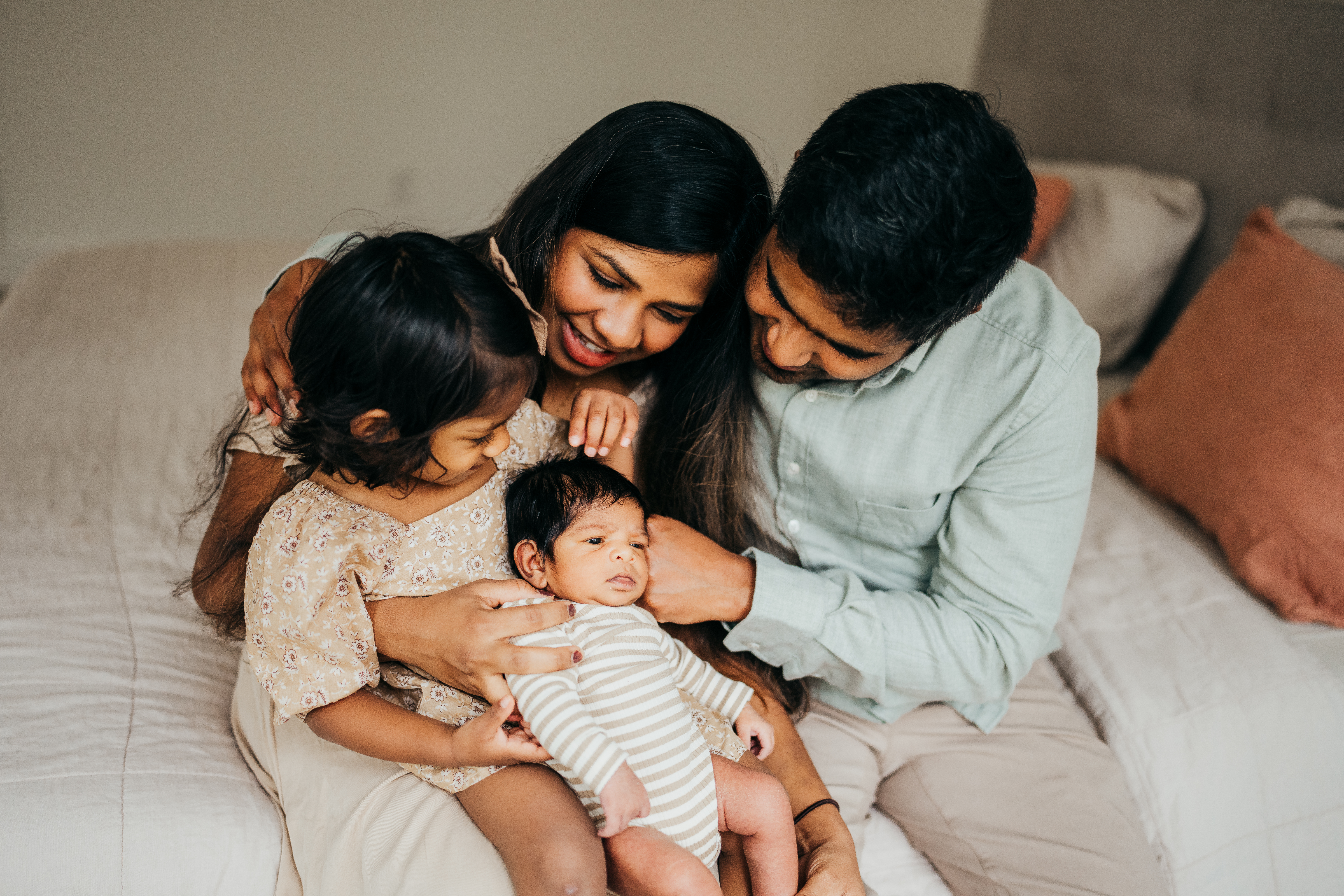 Happy mom and dad help their toddler daughter hold their newborn in her lap before visiting a daycare on mercer island