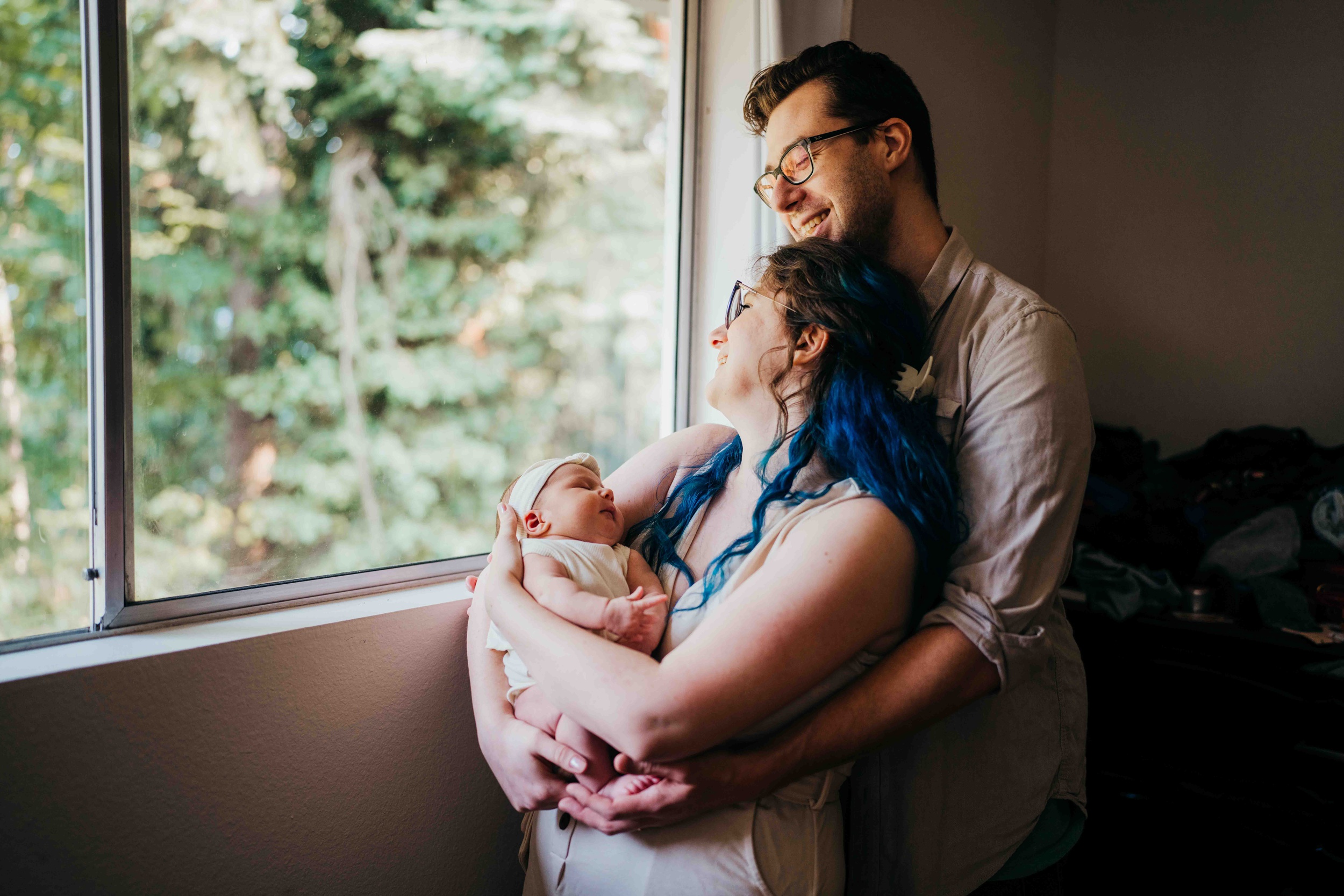 Happy new parents snuggle in a window holding their sleeping newborn baby before visiting fit4mom seattle