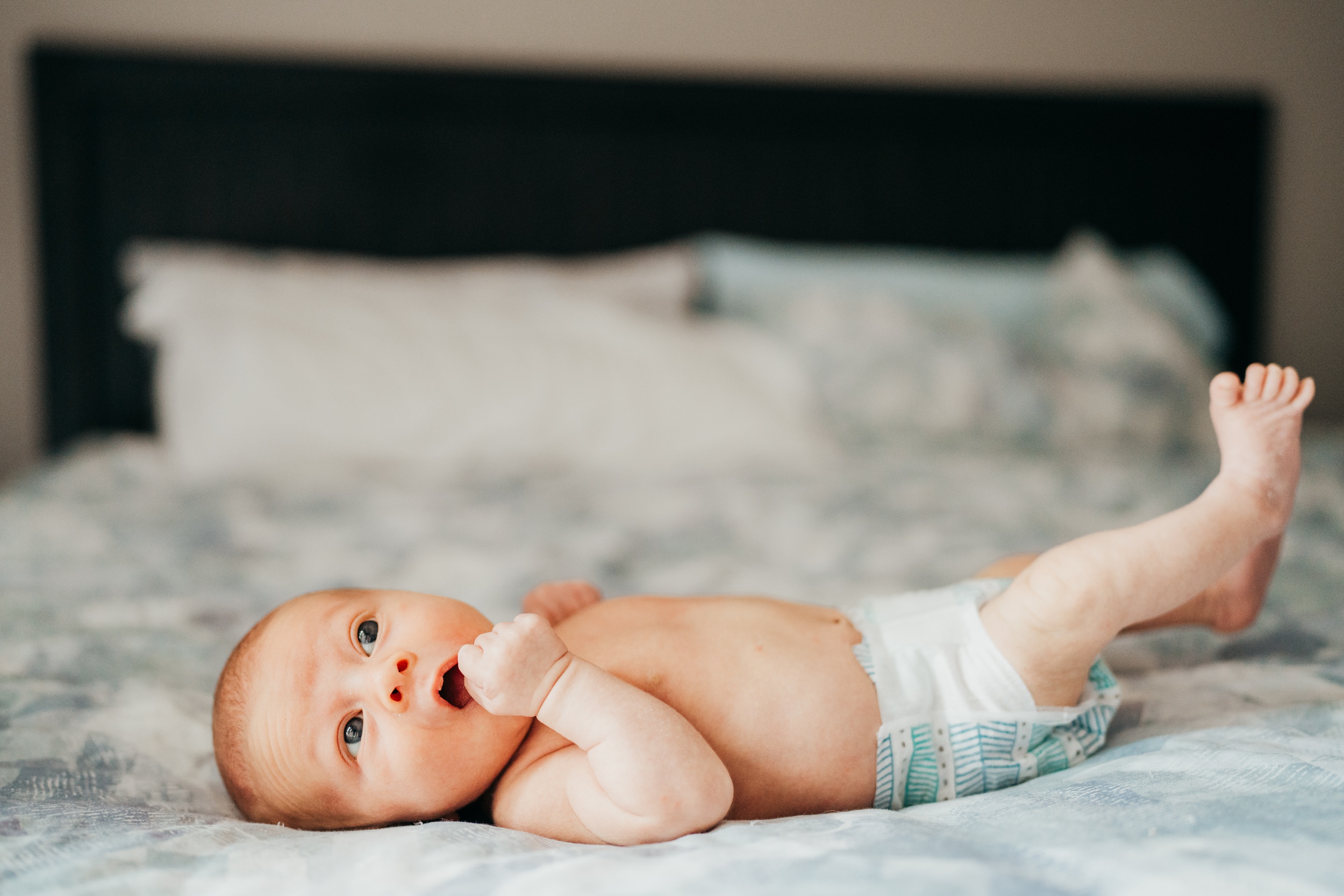 A newborn baby lays on a bed awake with a hand in its mouth