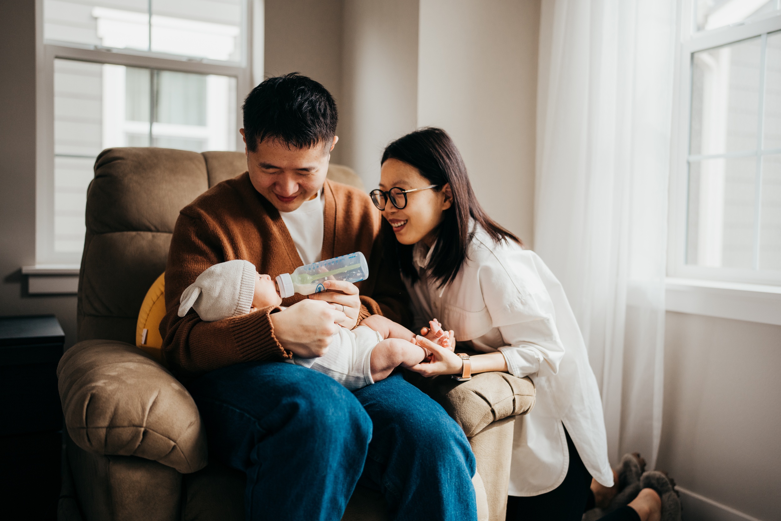 Happy new parents bottle feed their newborn baby in a chair