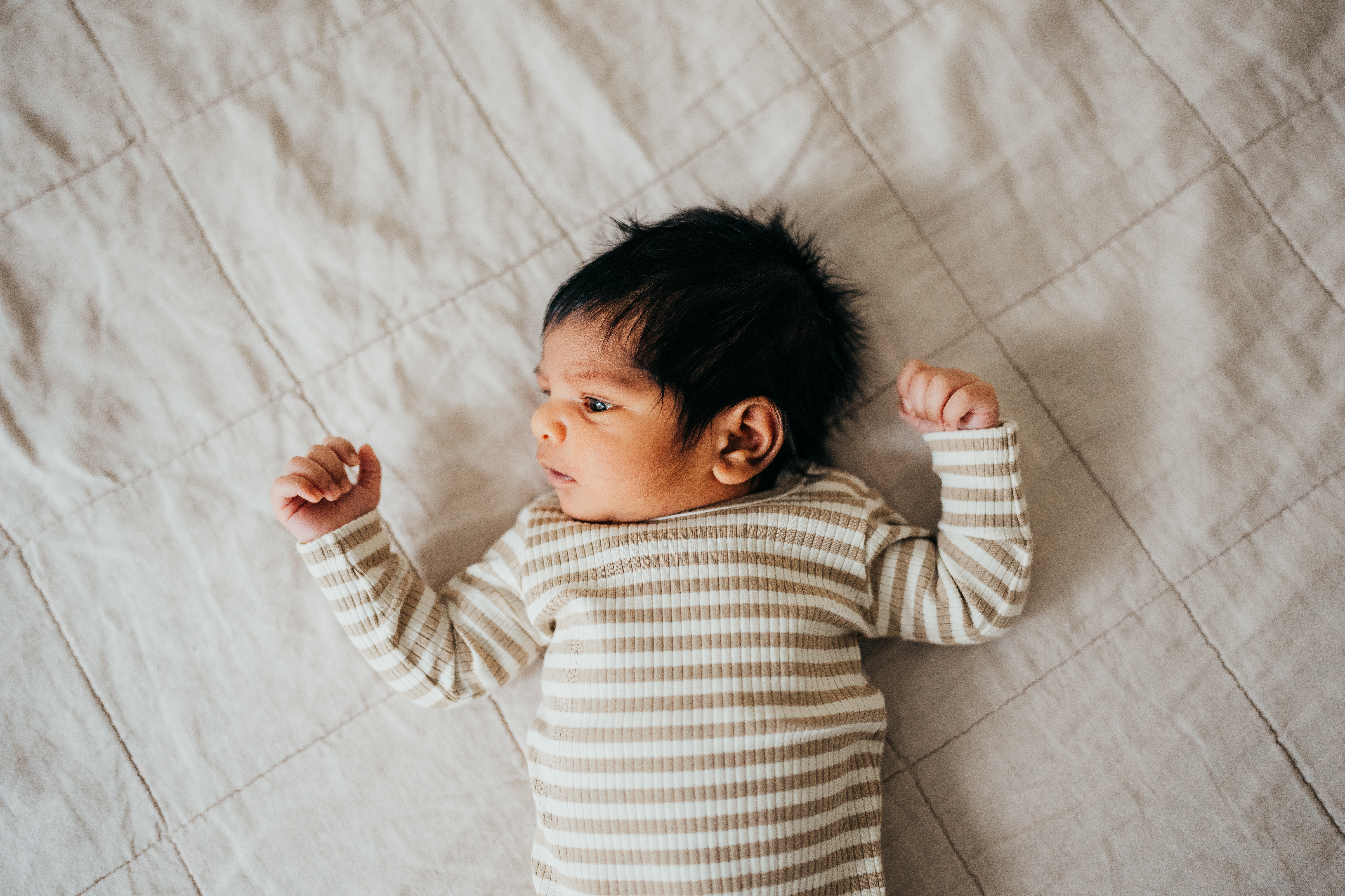 A newborn baby in a stripe onesie lays on a bed with eyes open looking at a window before visiting mercer island chiropractic