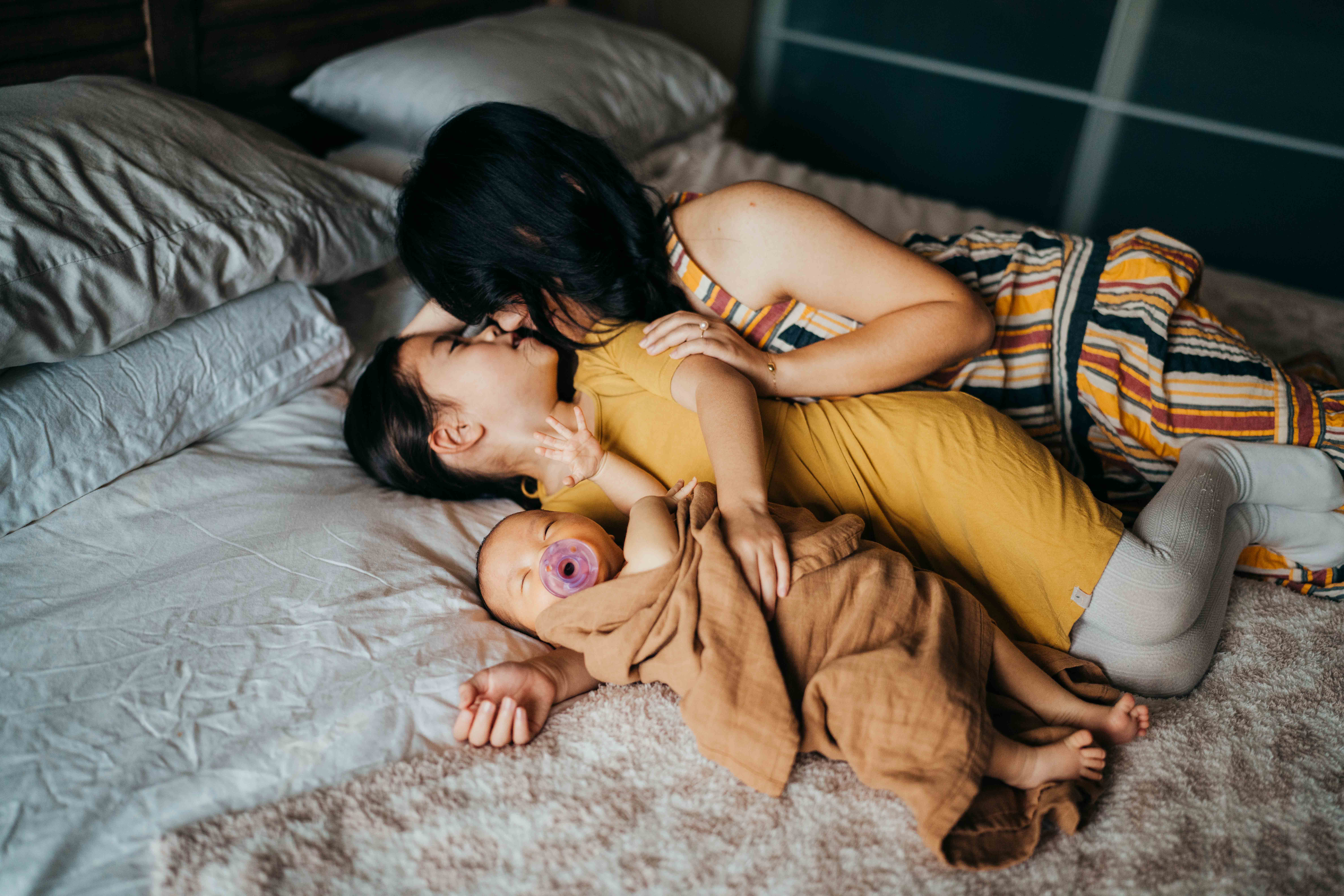 A mom snuggles her toddler daughter and newborn baby on a bed