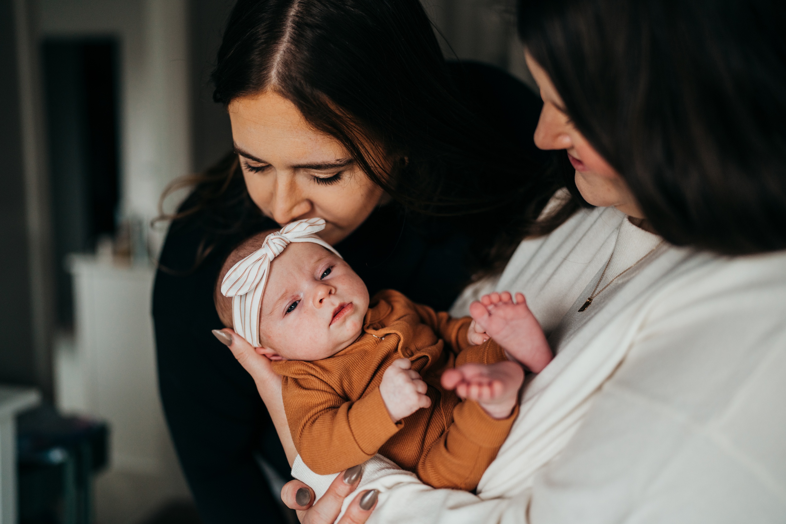 Happy moms kiss and admire their newborn in their arms in an orange onesie and white headband before some mommy and me classes in seattle