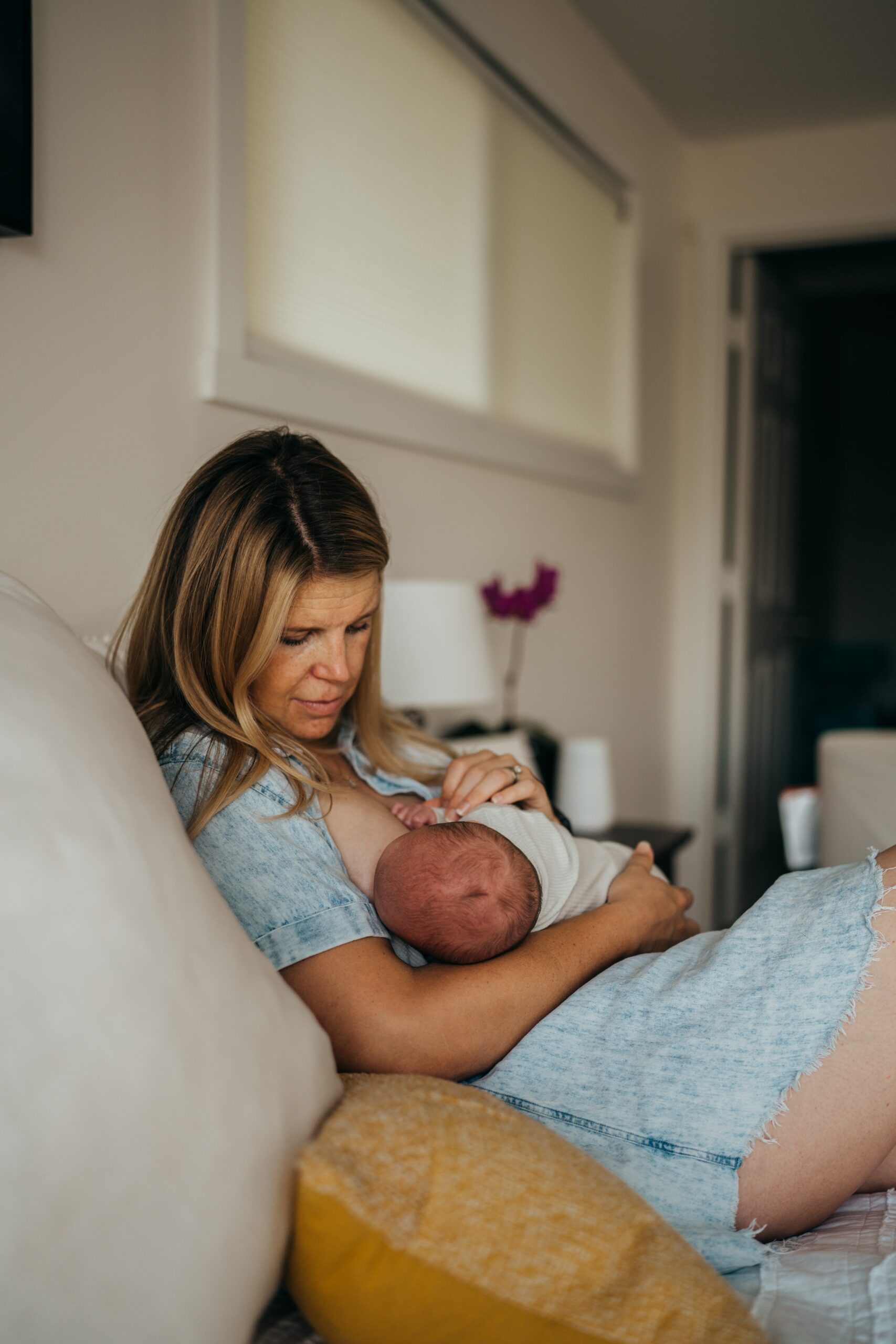 A mom breastfeeds her newborn baby on a leather couch in a denim dress