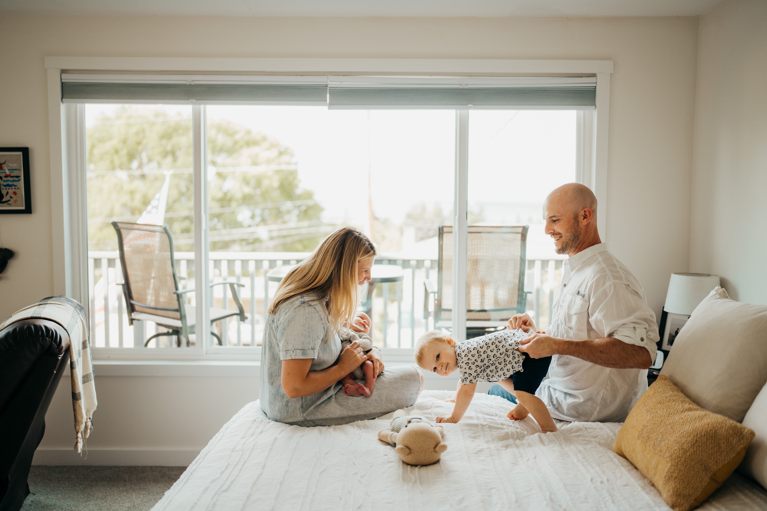 Happy mom and dad play with their toddler son no a bed while their newborn sleeps in mom's lap thanks to an obgyn in seattle