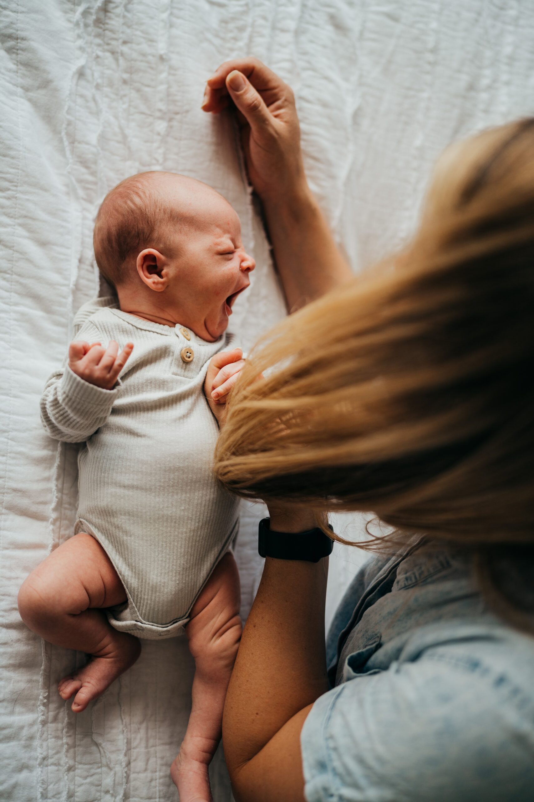 A newborn baby yawns while laying on a bed under mom in a white onesie thanks to an obgyn in seattle