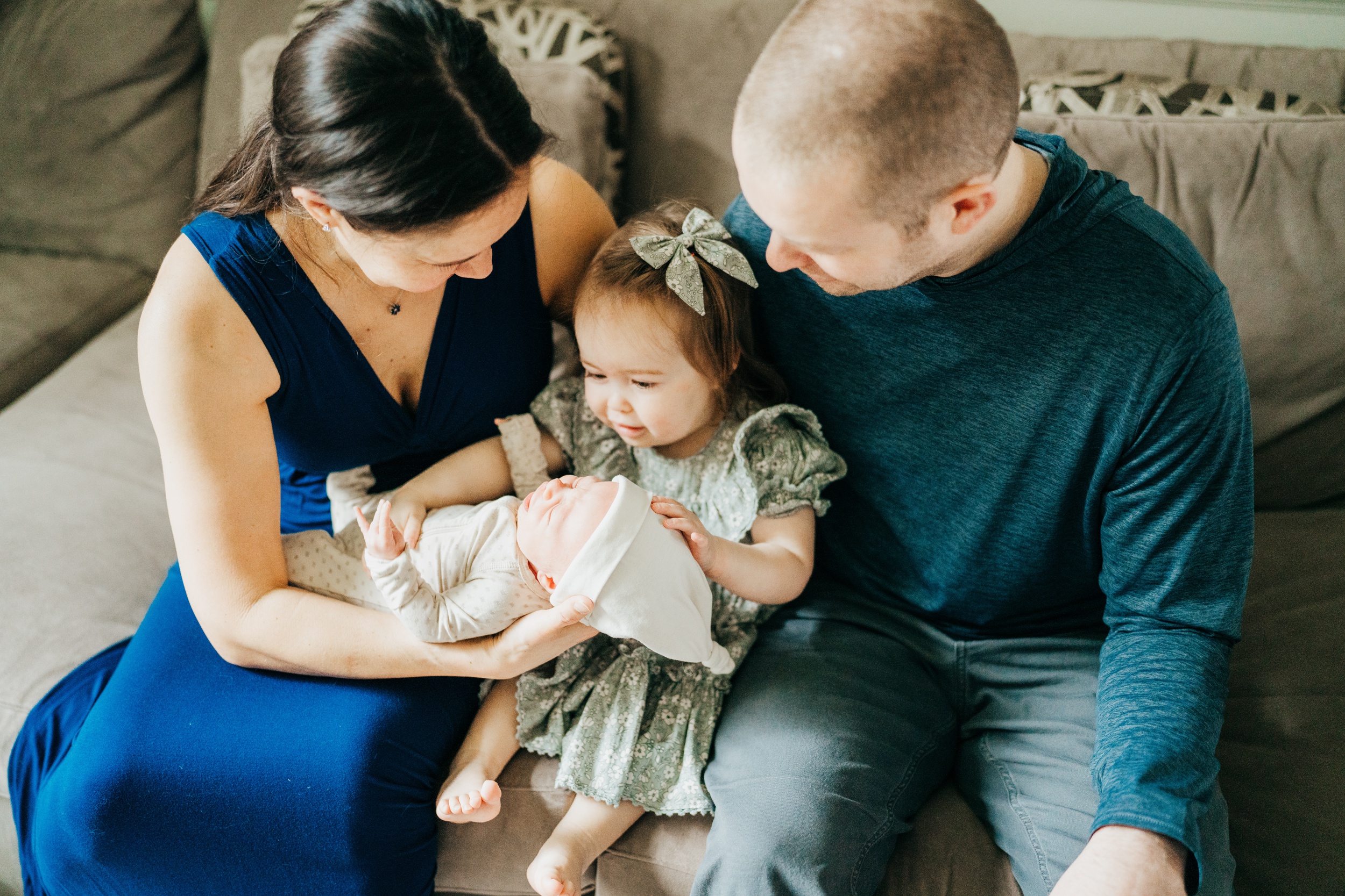 A toddler girl in a green dress reaches for her sleeping newborn baby sibling in mom's hands as they sit on a couch with dad