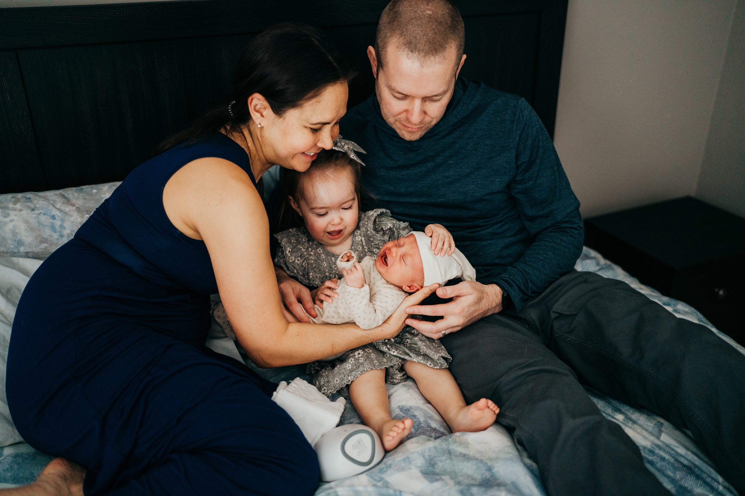 Happy new parents help their toddler daughter hold their newborn while sitting on a bed after parenting classes in seattle