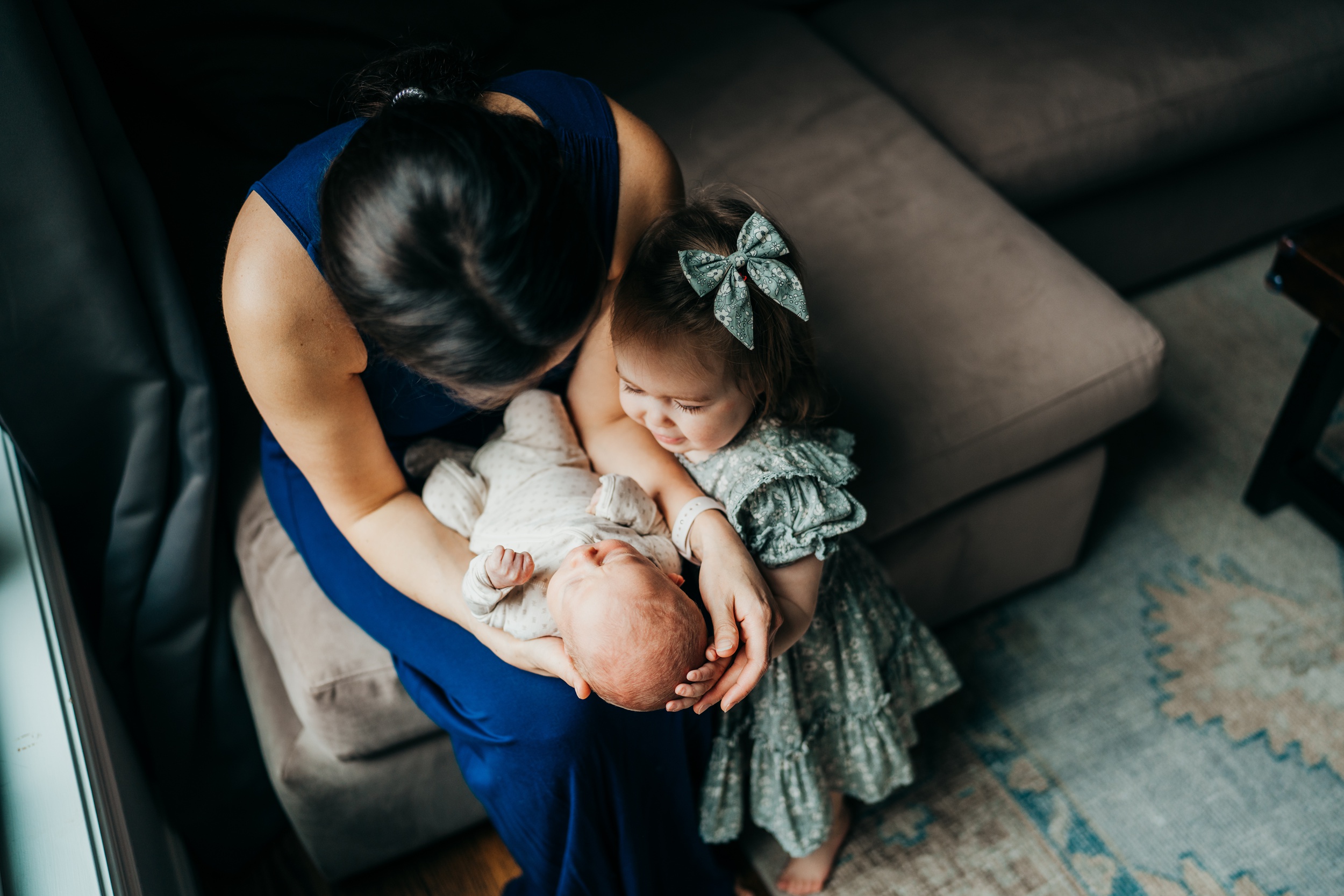A toddler girl admires her sleeping newborn sibling in mom's lap on a couch thanks to parenting classes in seattle