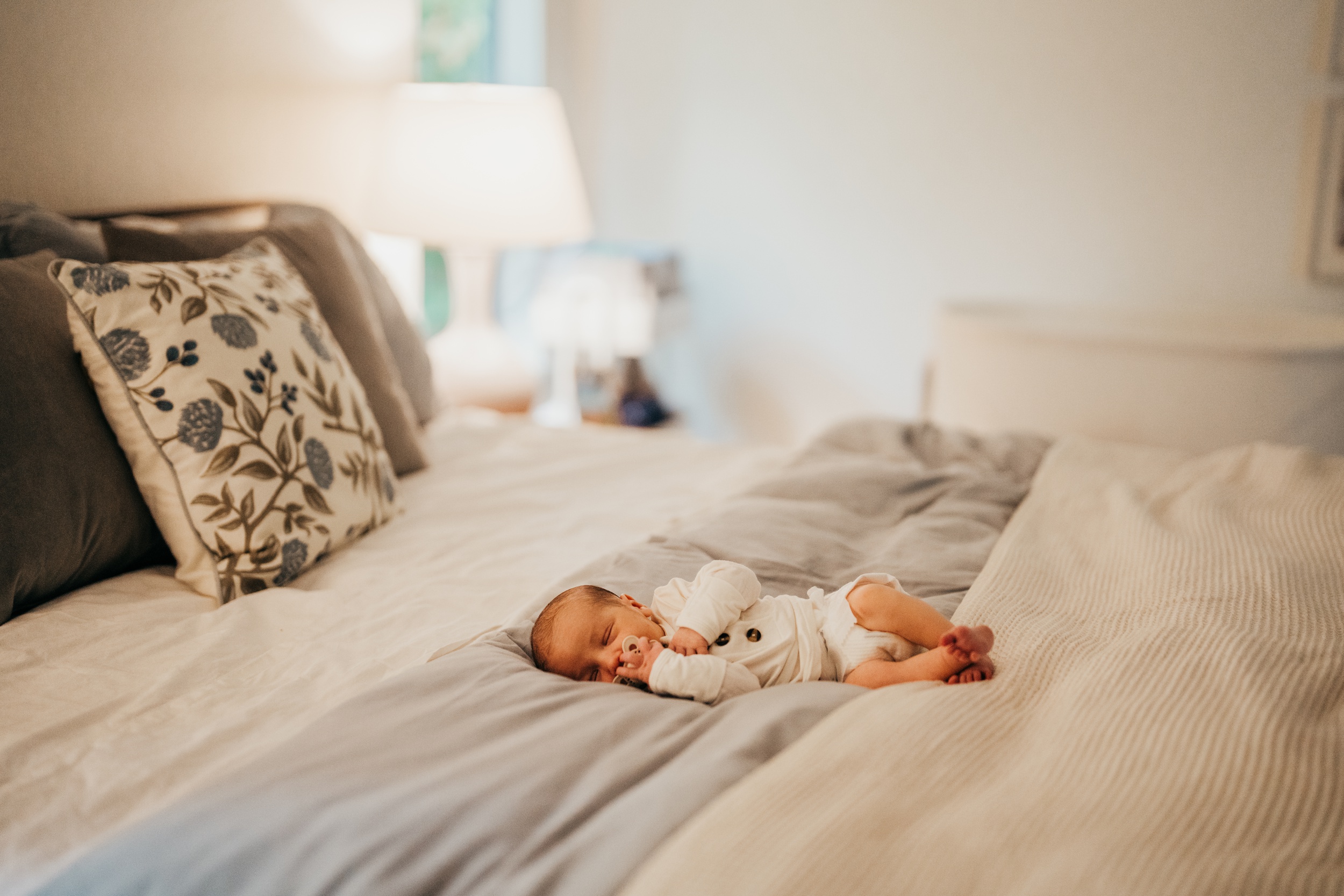 A newborn abby in a white onesie sleeps with a binky on a bed