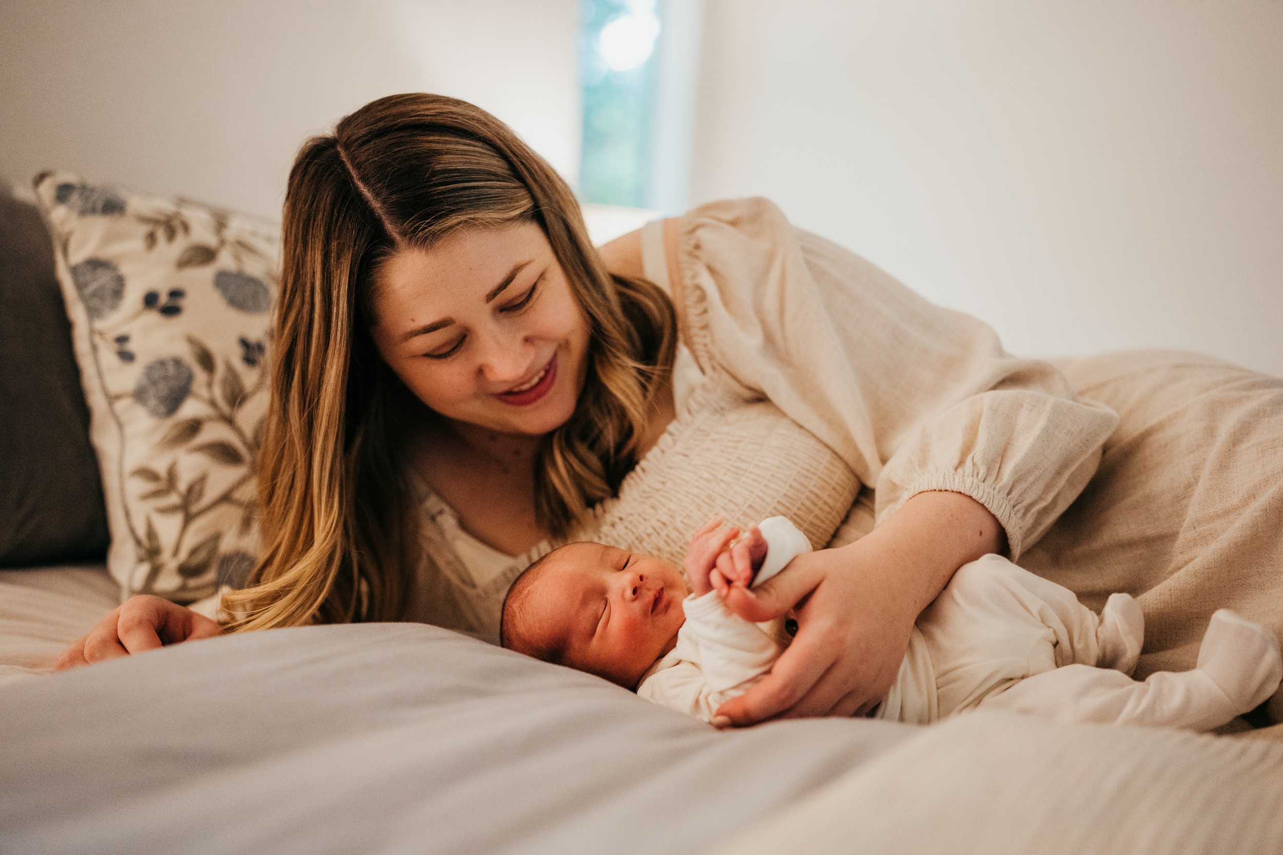 A smiling new mom plays with her sleeping newborn on a bed in a cream dress