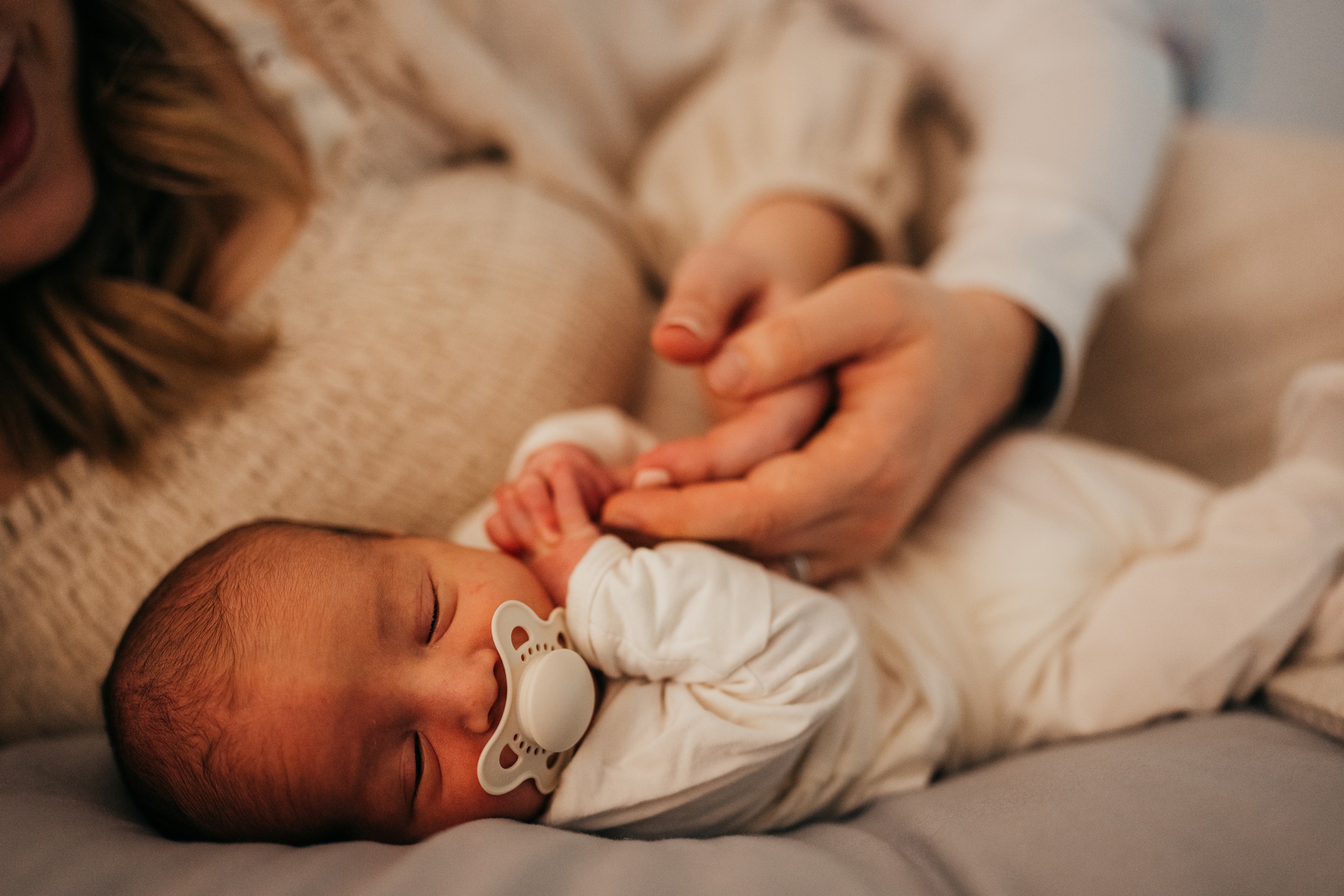 A newborn baby sleeps in a white onesie while holding mom and dad's fingers after finding placenta encapsulation in seattle