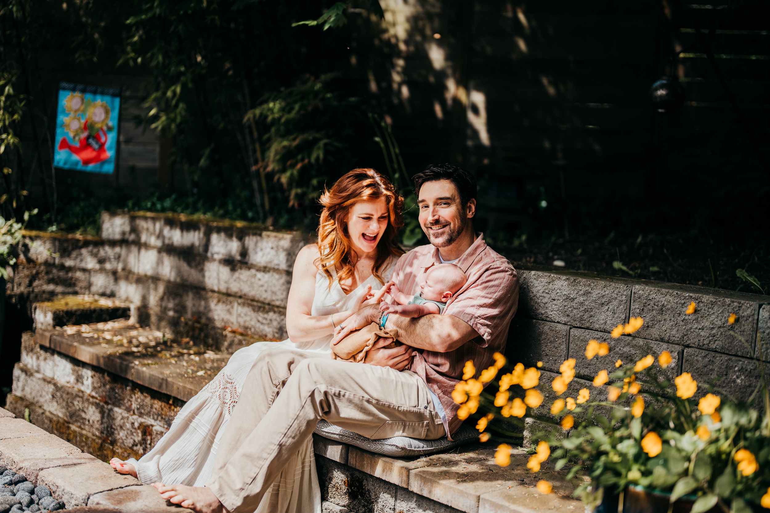 A laughing mom plays with her newborn laying in dad's lap in a garden
