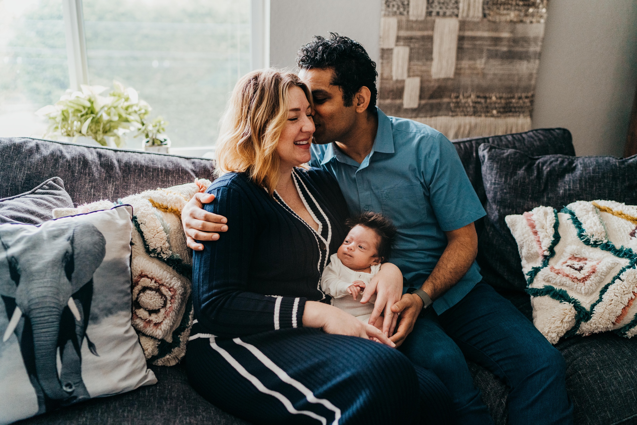A new mom smiles while dad kisses her cheek with newborn awake in her lap