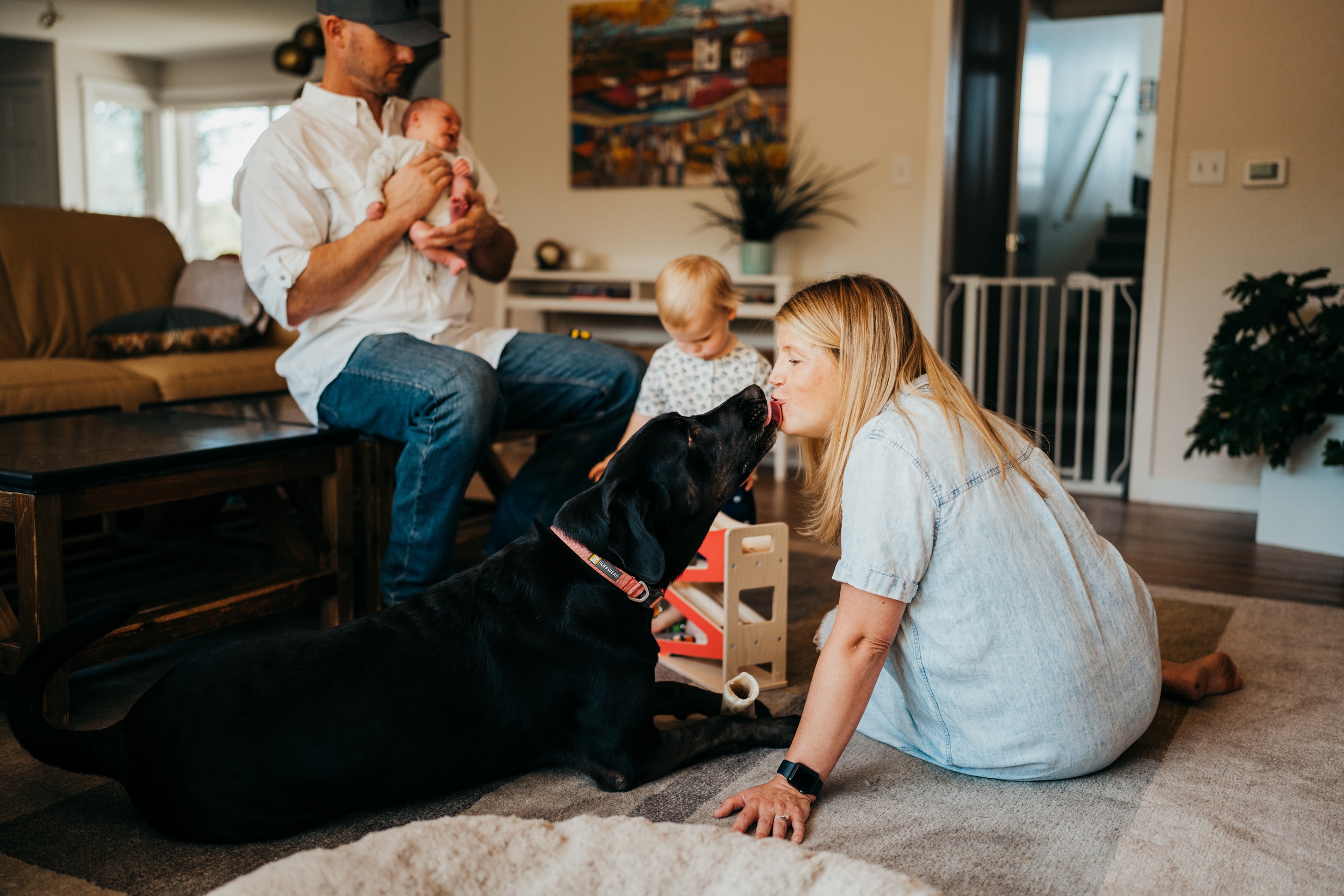 A black dog kisses mom sitting on the floor while dad holds a newborn and toddler plays with wooden toys before some swim lessons on mercer island