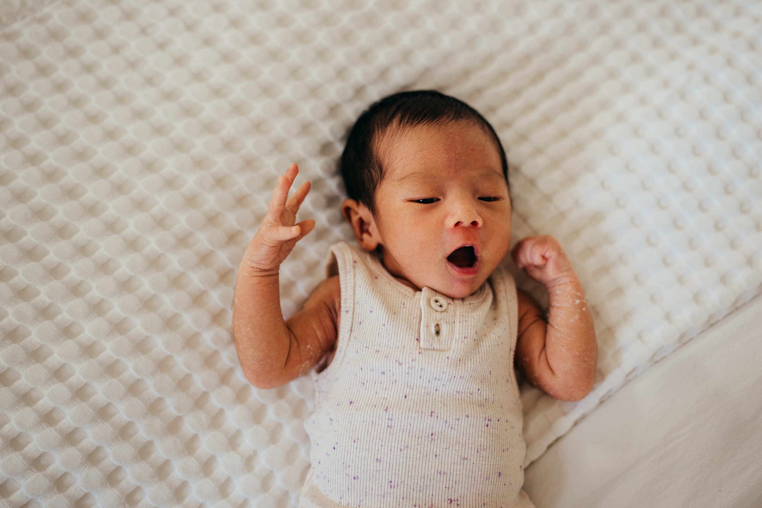 A newborn baby yawns while laying on a bed in a sleeveless onesie