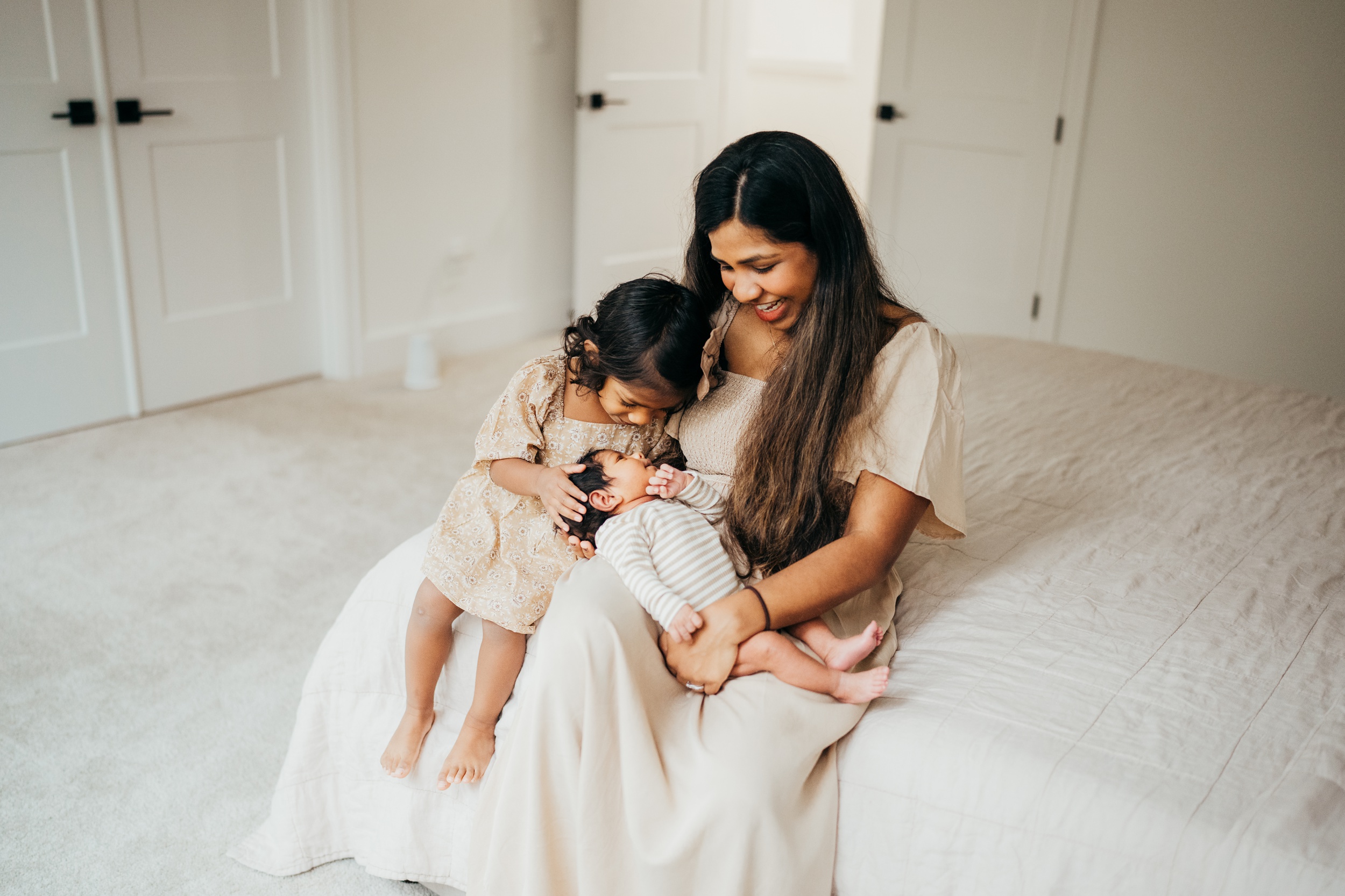 A toddler girl smiles over her newborn sibling in mom's lap on a bed after visiting thistle and poppy