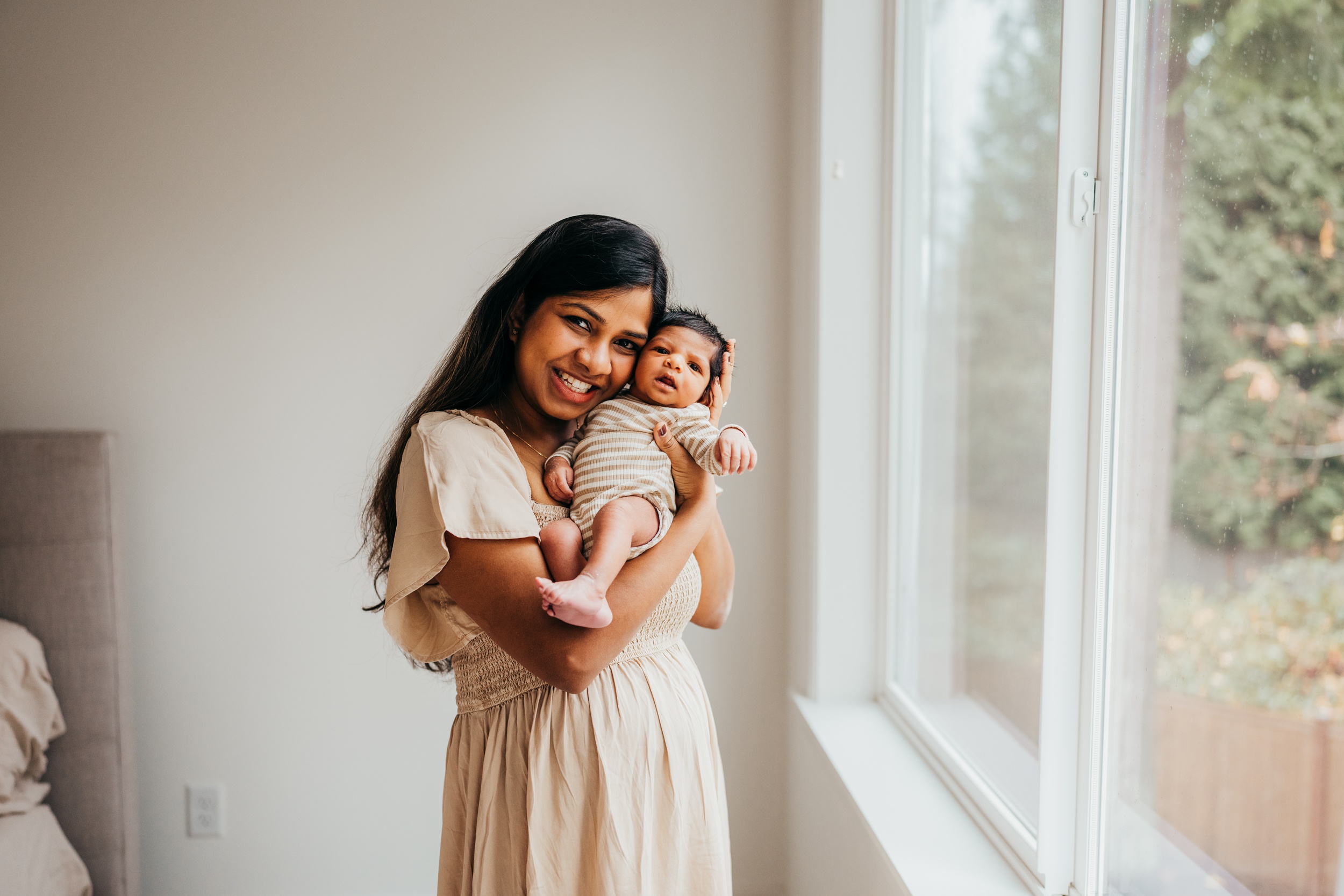 A joyful mom stands in a bedroom window cradling her awake newborn against her cheek in a cream dress after visiting thistle and poppy