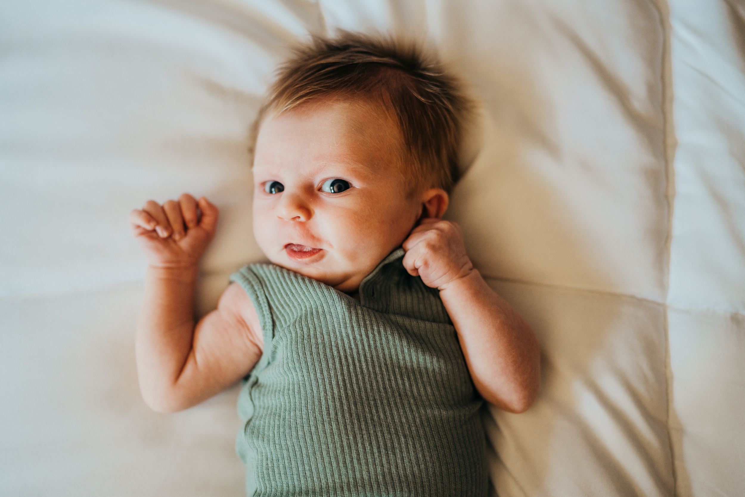 A newborn baby with eyes open in a green onesie lays on a bed