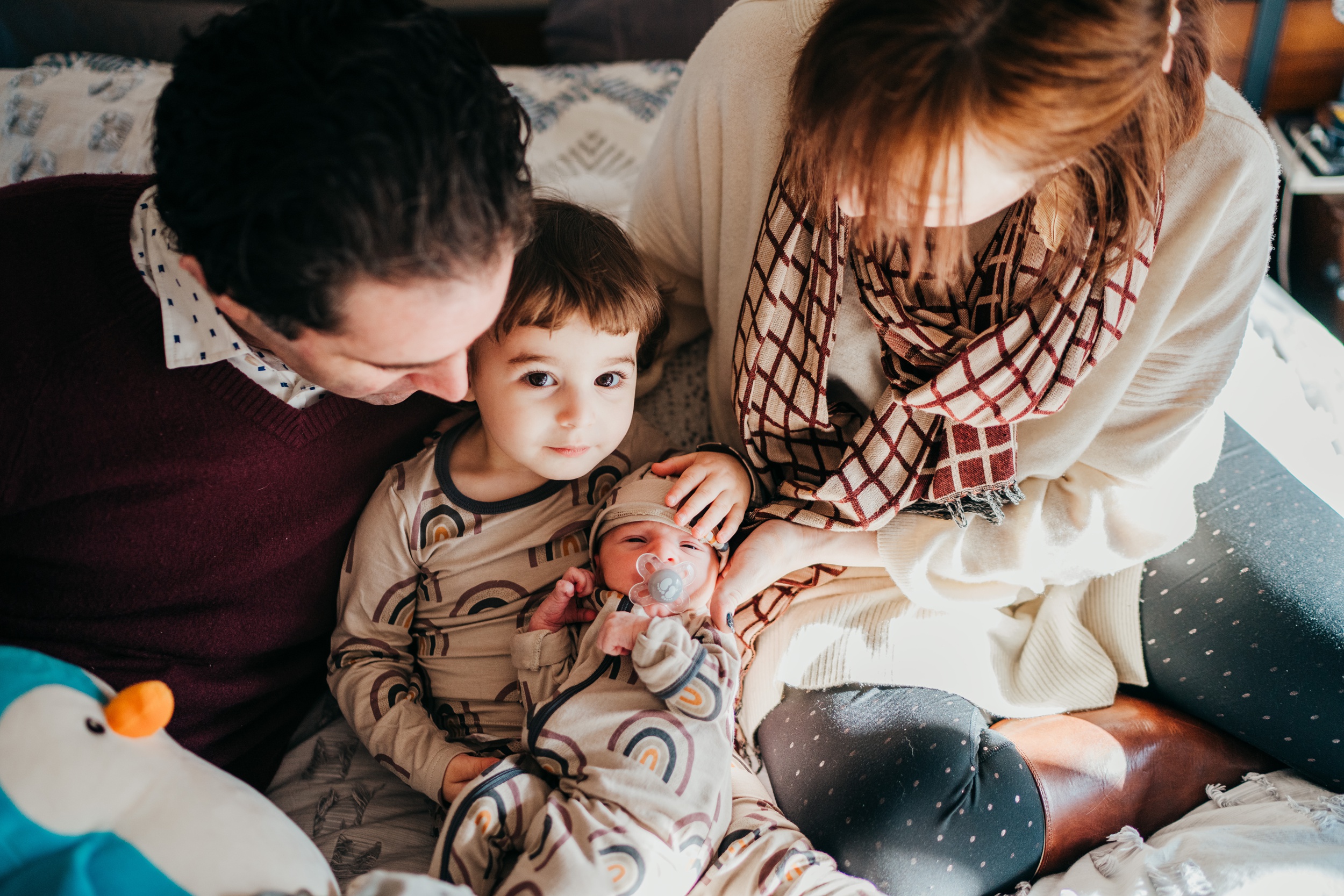 A mom and dad cuddle their toddler and newborn in matching rainbow pajamas on a bed