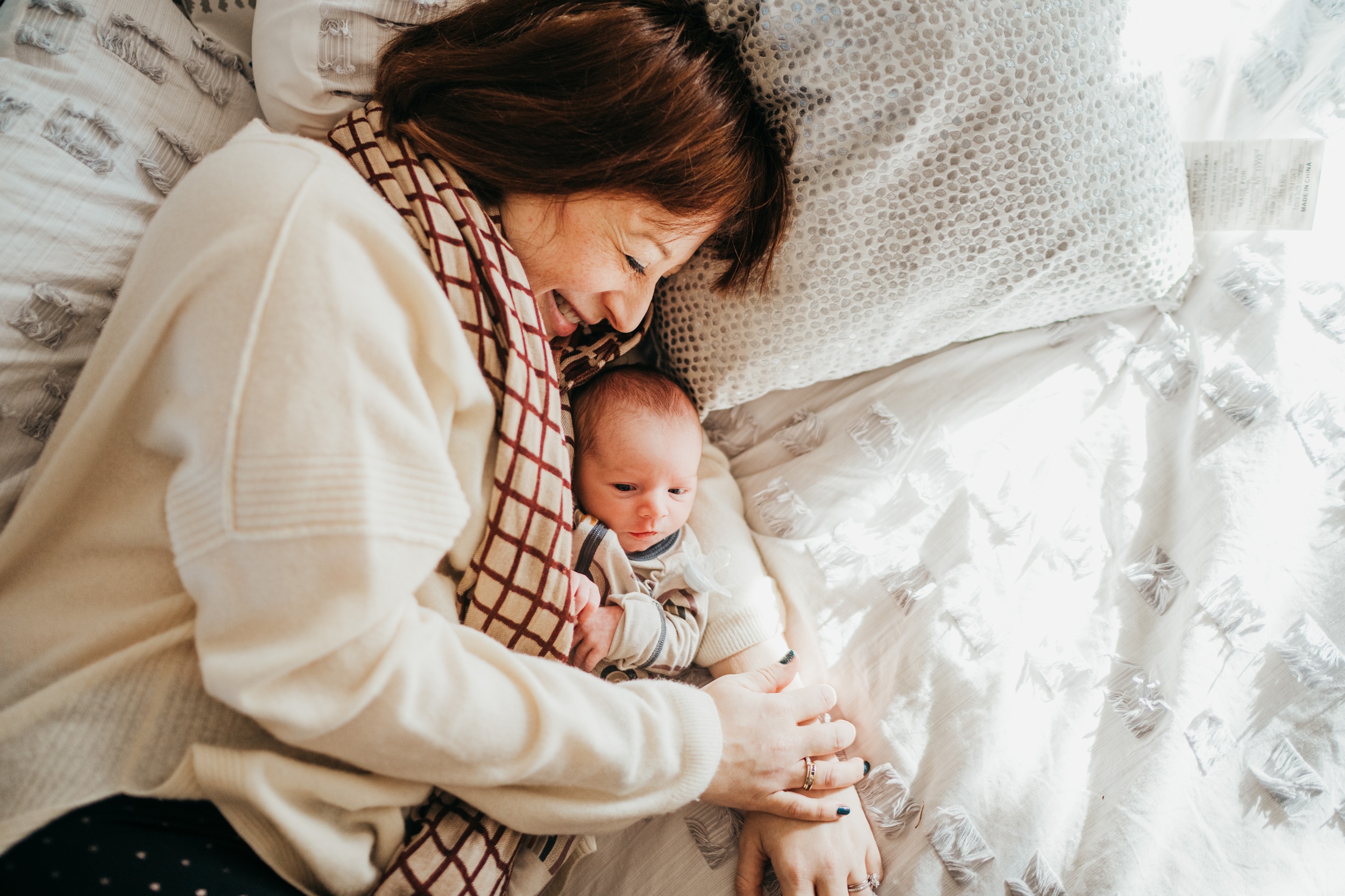 A happy mom in a white sweater and red scarf snuggles her newborn on a bed after visiting bootyland kids