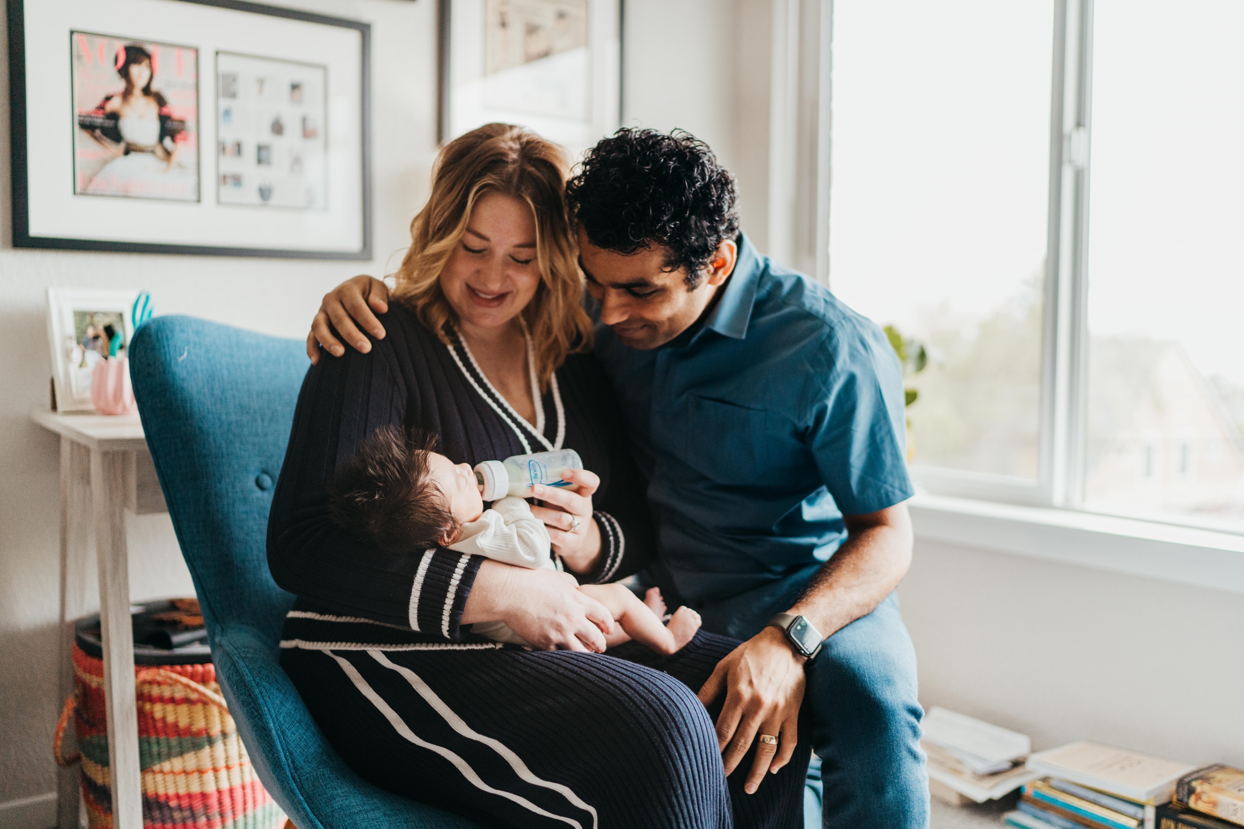 Smiling mom and dad feed their newborn with a bottle while sitting in a blue chair under a window after using city mouse studio