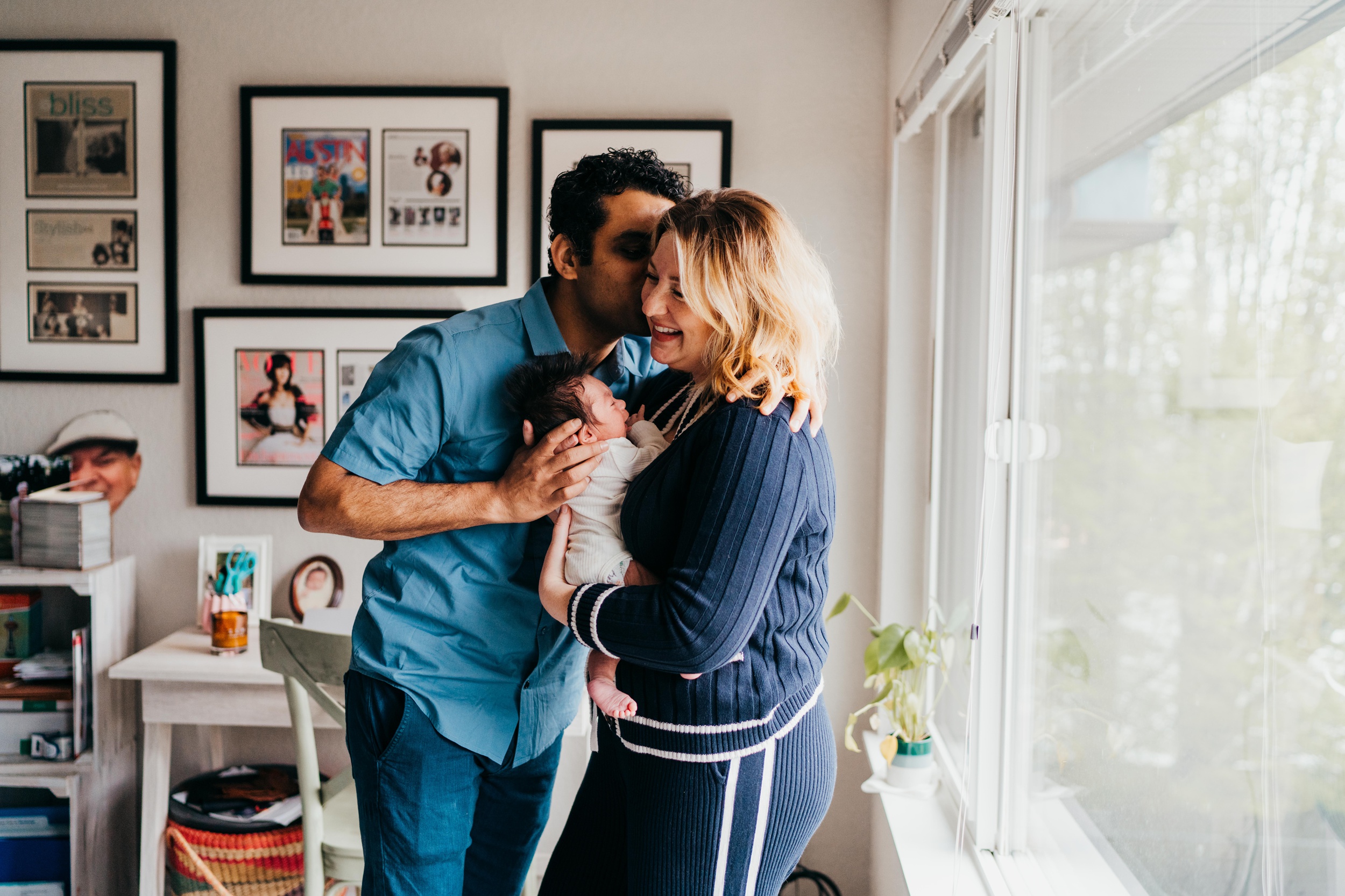 Happy new parents laugh and kiss while holding their newborn in a window in blue after using city mouse studio
