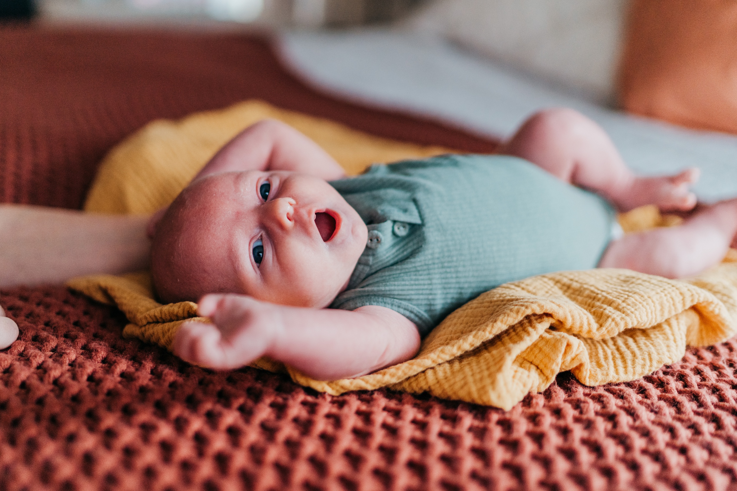 A newborn baby reaches above his head while laying on a bed in a blue onesie