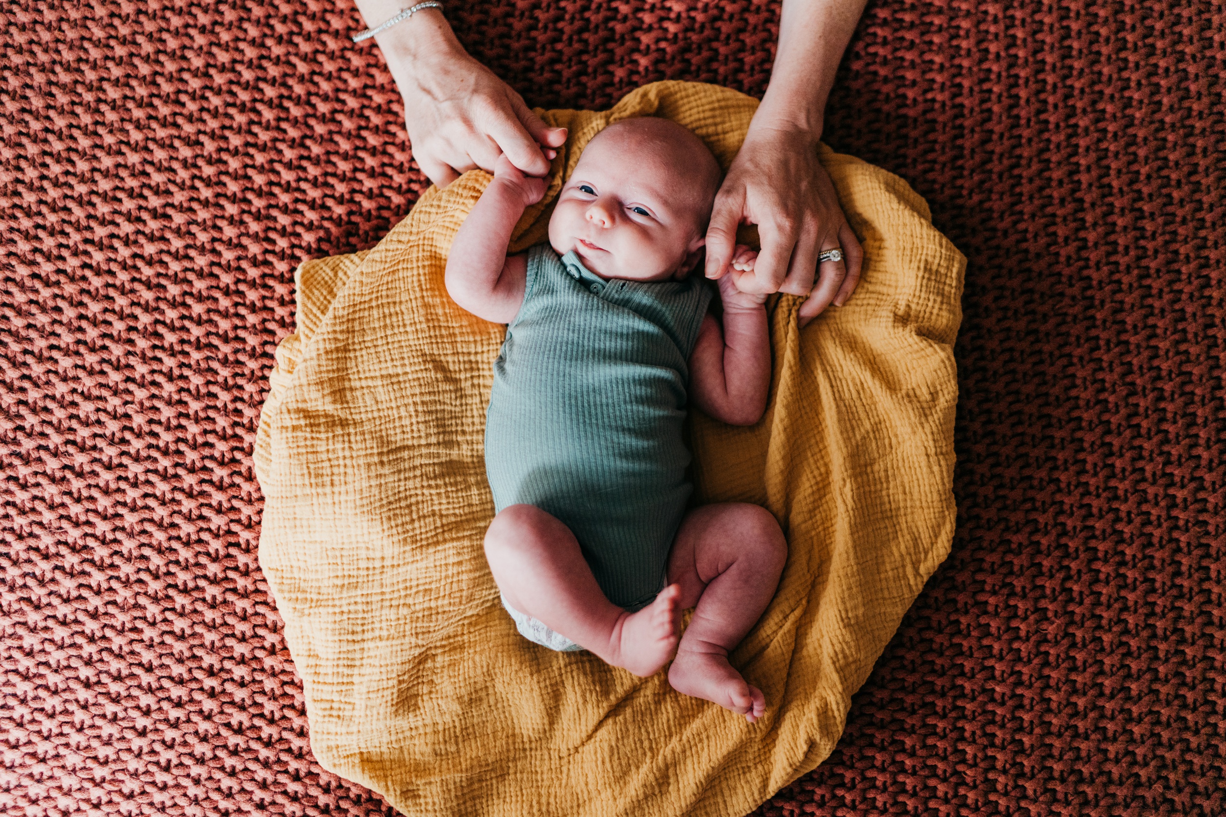 A happy newborn lays on a yellow blanket on a bed while holding mom's fingers after visiting clover toys