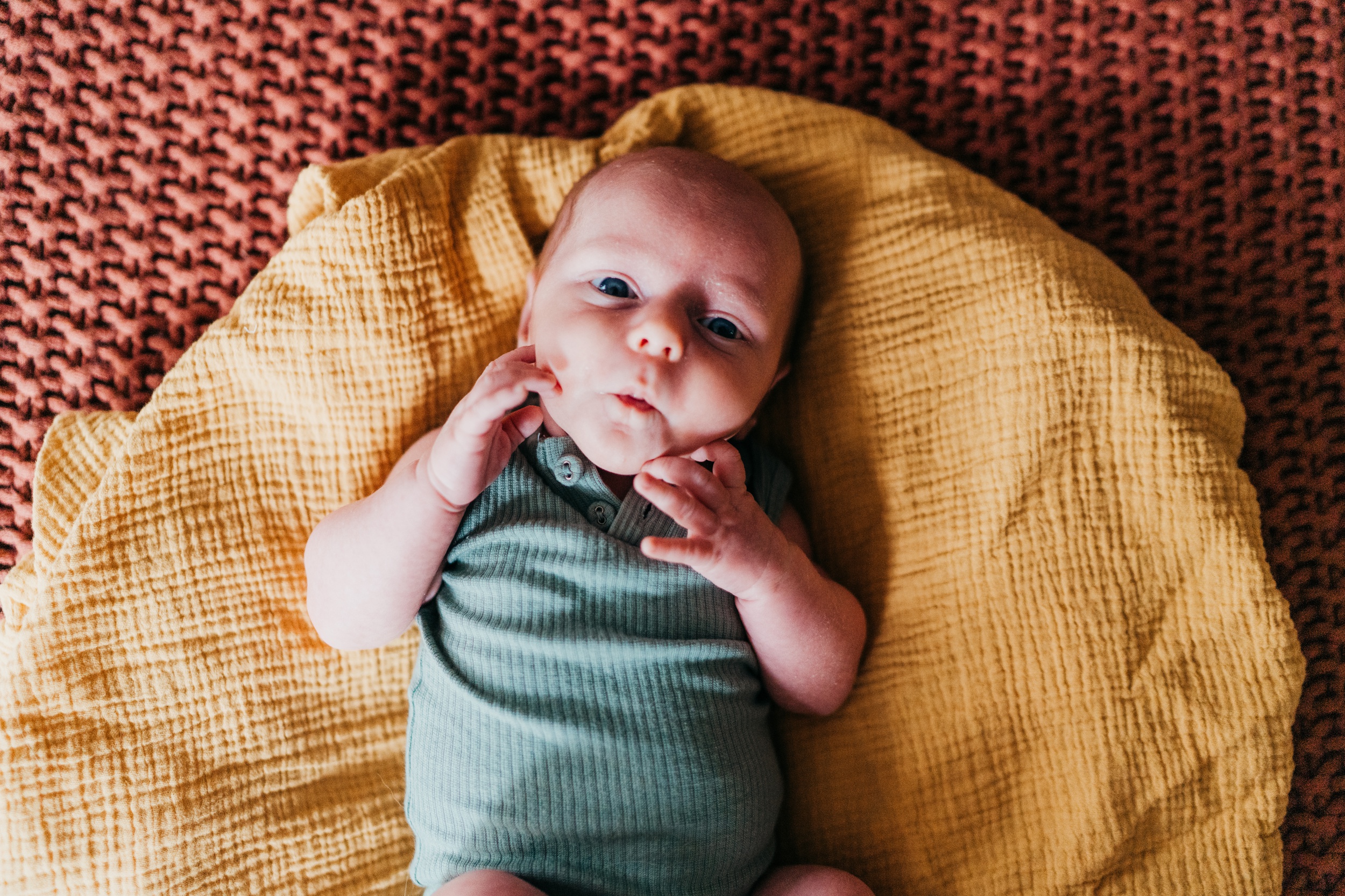 A newborn baby makes a silly face while laying on a yellow blanket in a blue onesie after visiting clover toys