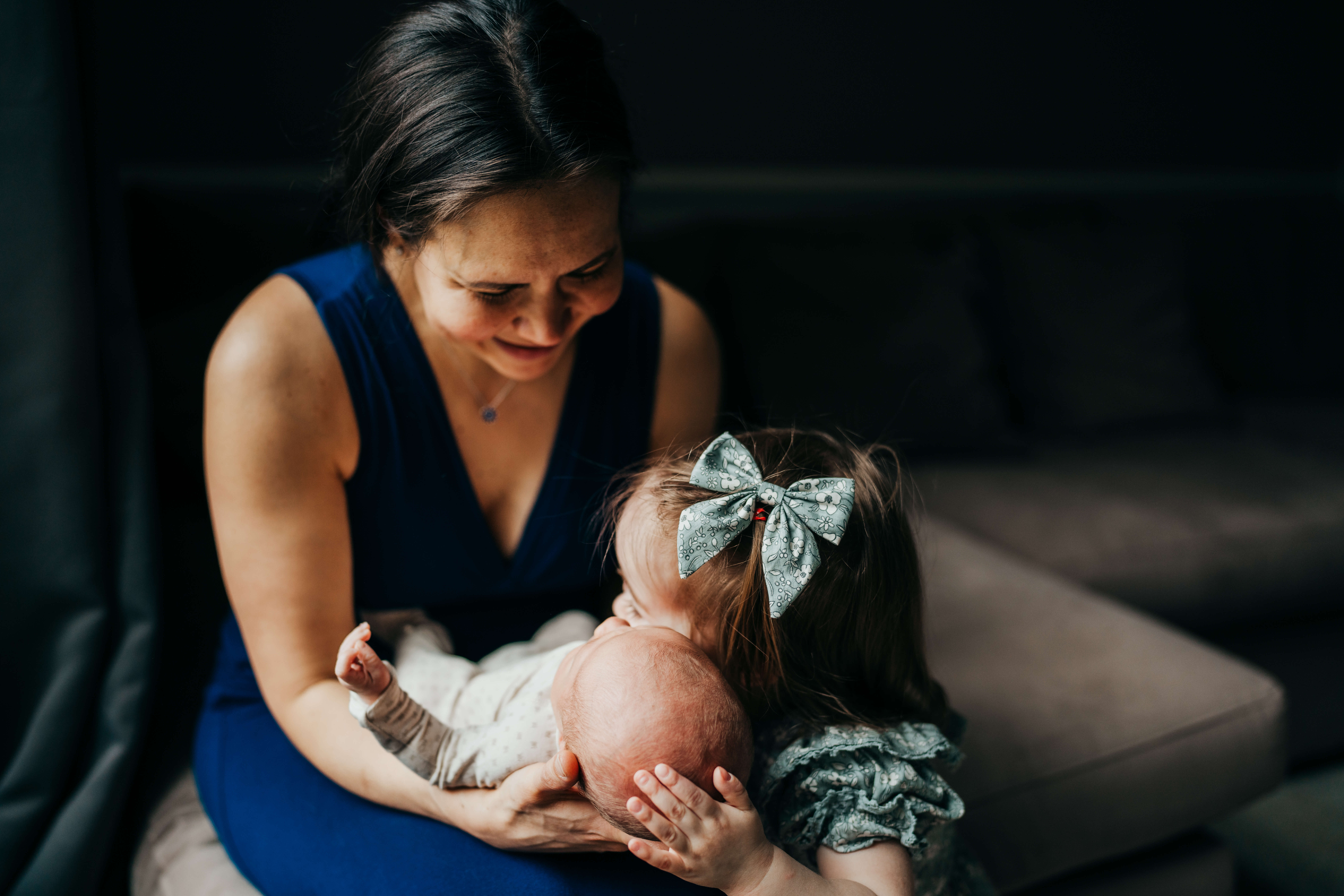 a mother in a blue dress smiles while watching her toddler hug and touch cheeks with newborn in her hands after leaving Puget Sound Birth Center