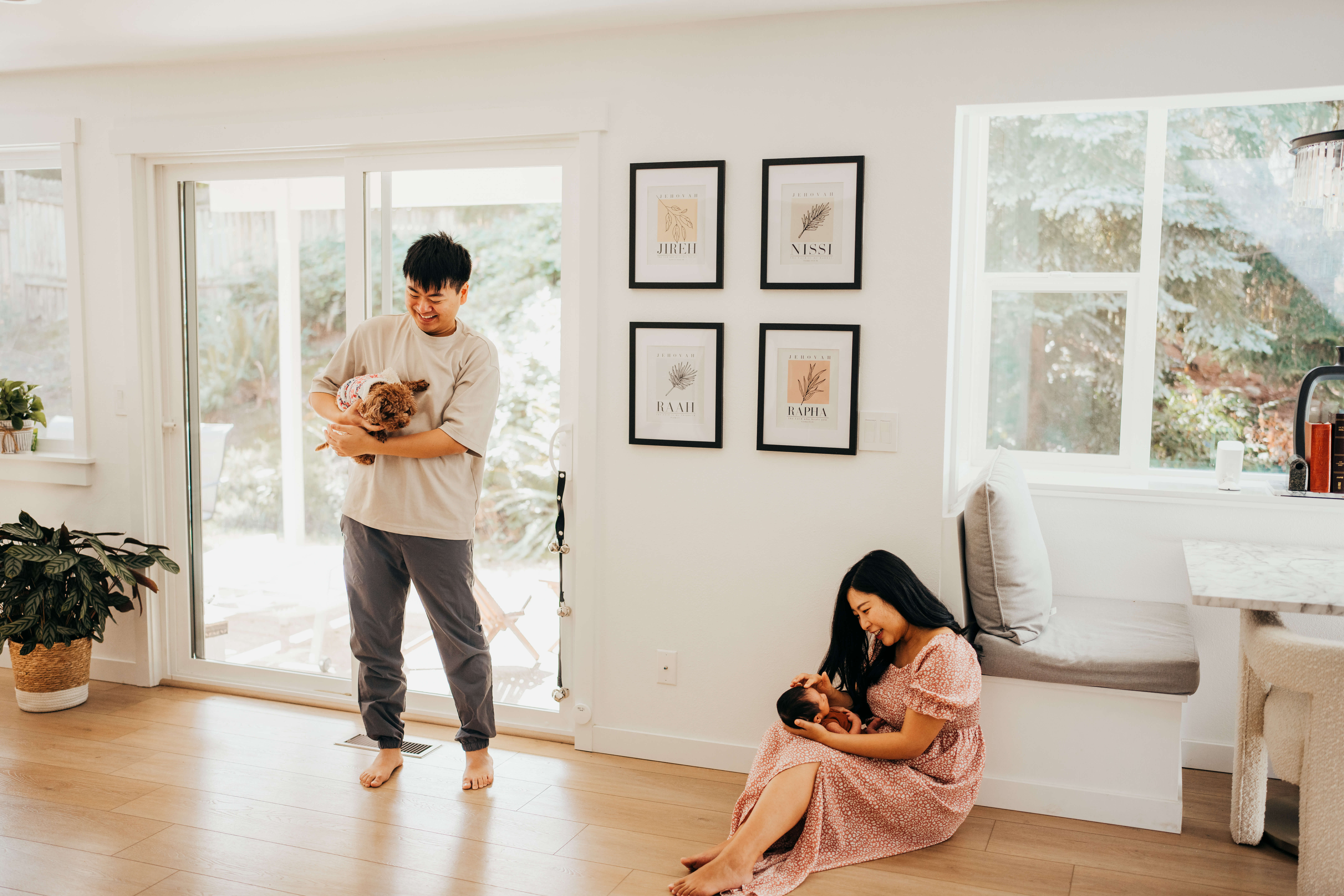 a mom sits ont he floor of their kitchen playing with their newborn as dad stands in a doorway playing with their tiny puppy