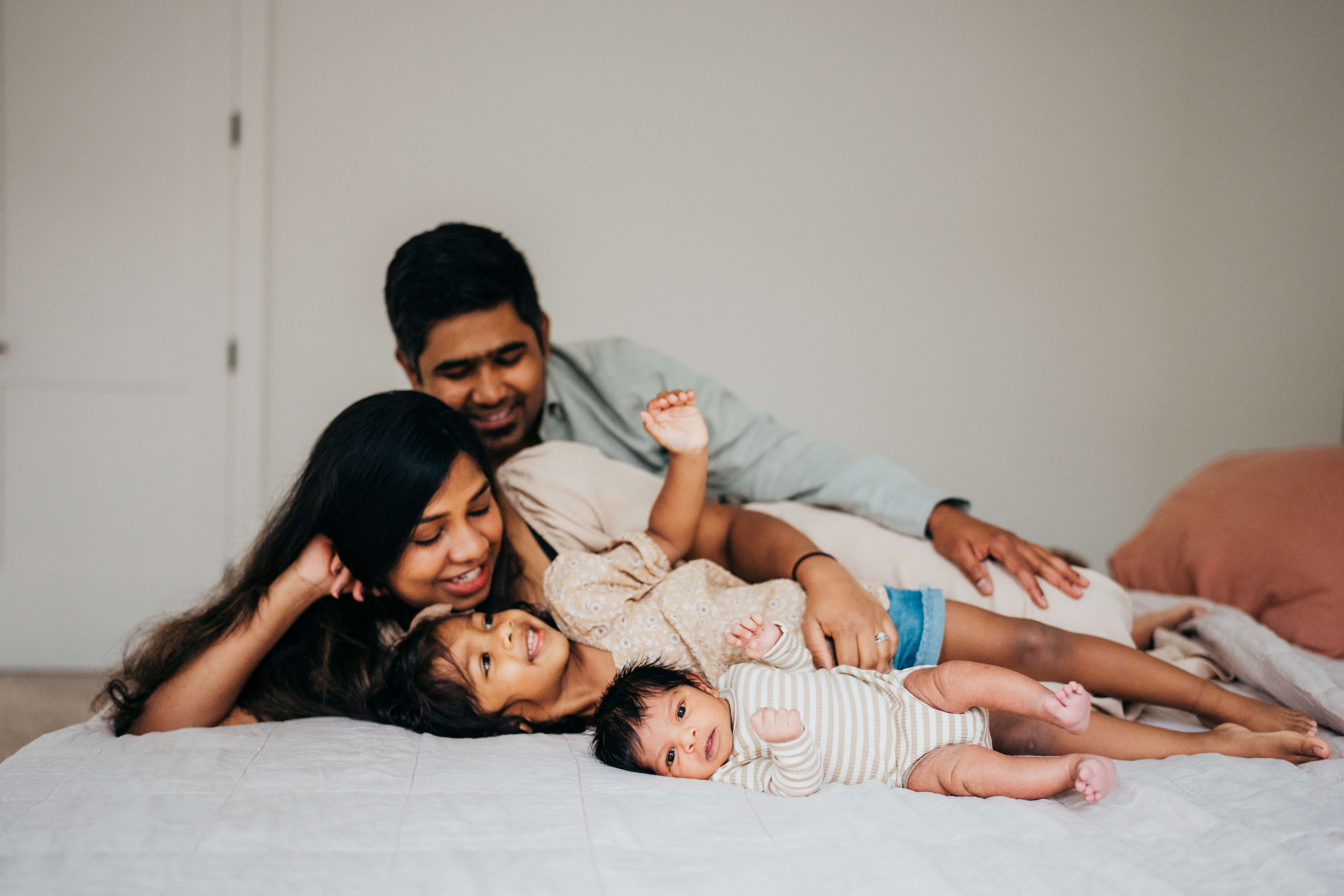 A mom and dad lay on a bed with their toddler daughter and newborn as they all laugh after meeting an au pair in seattle