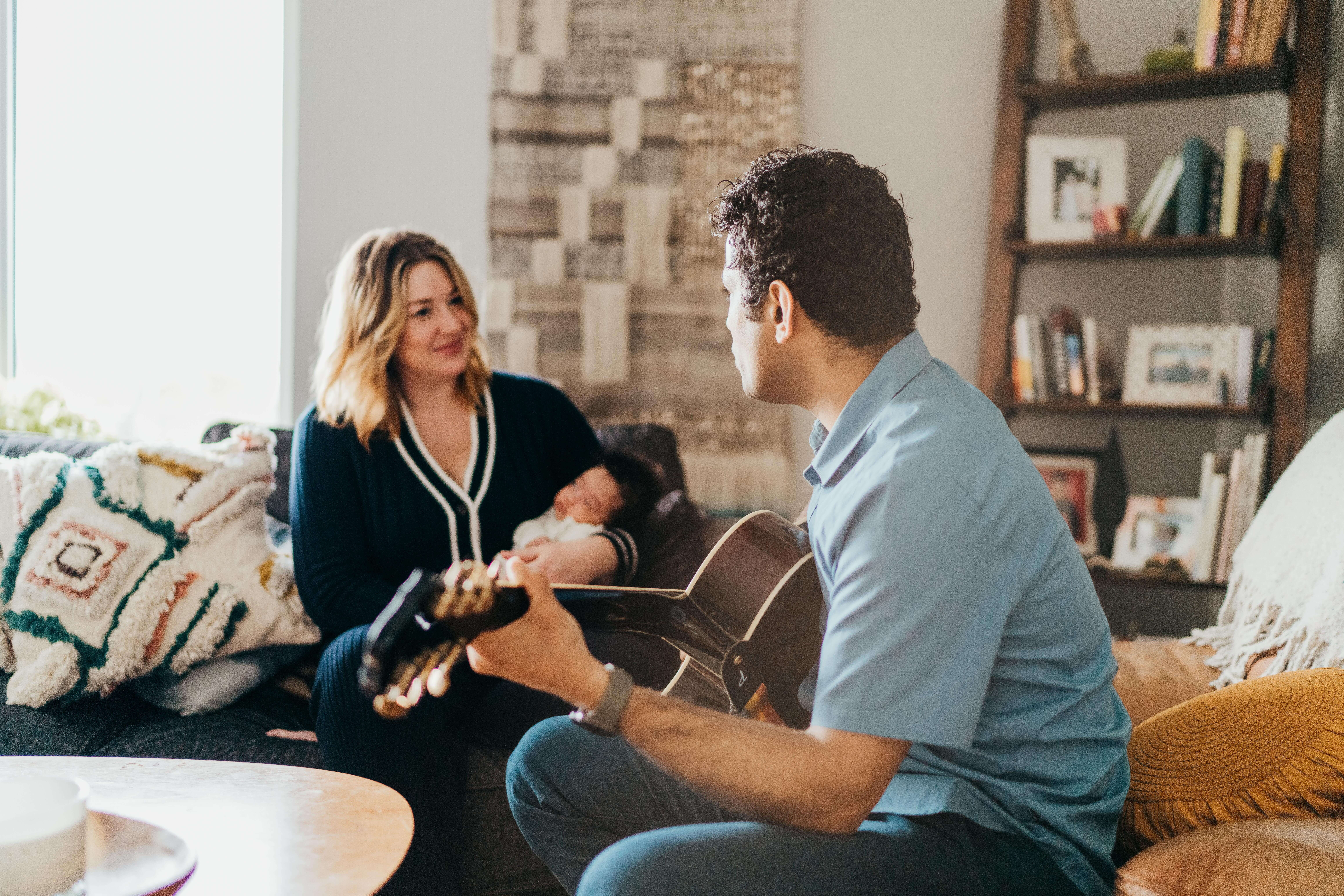 A dad sits on a couch playing guitar while mom smiles at him with their newborn in her arms
