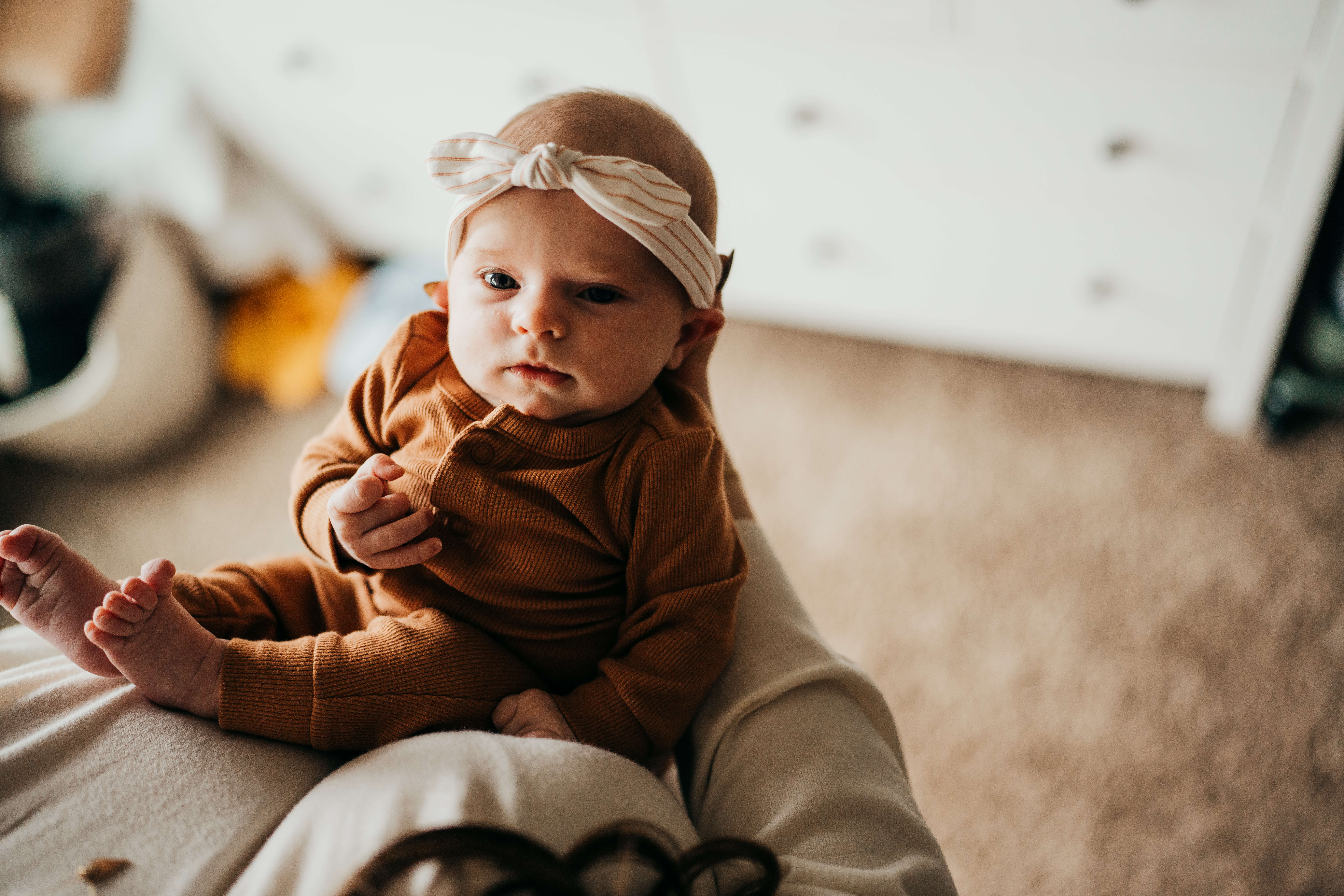 a newborn baby in a headband and brown onesie sits in mom's hands after visiting flora and henri