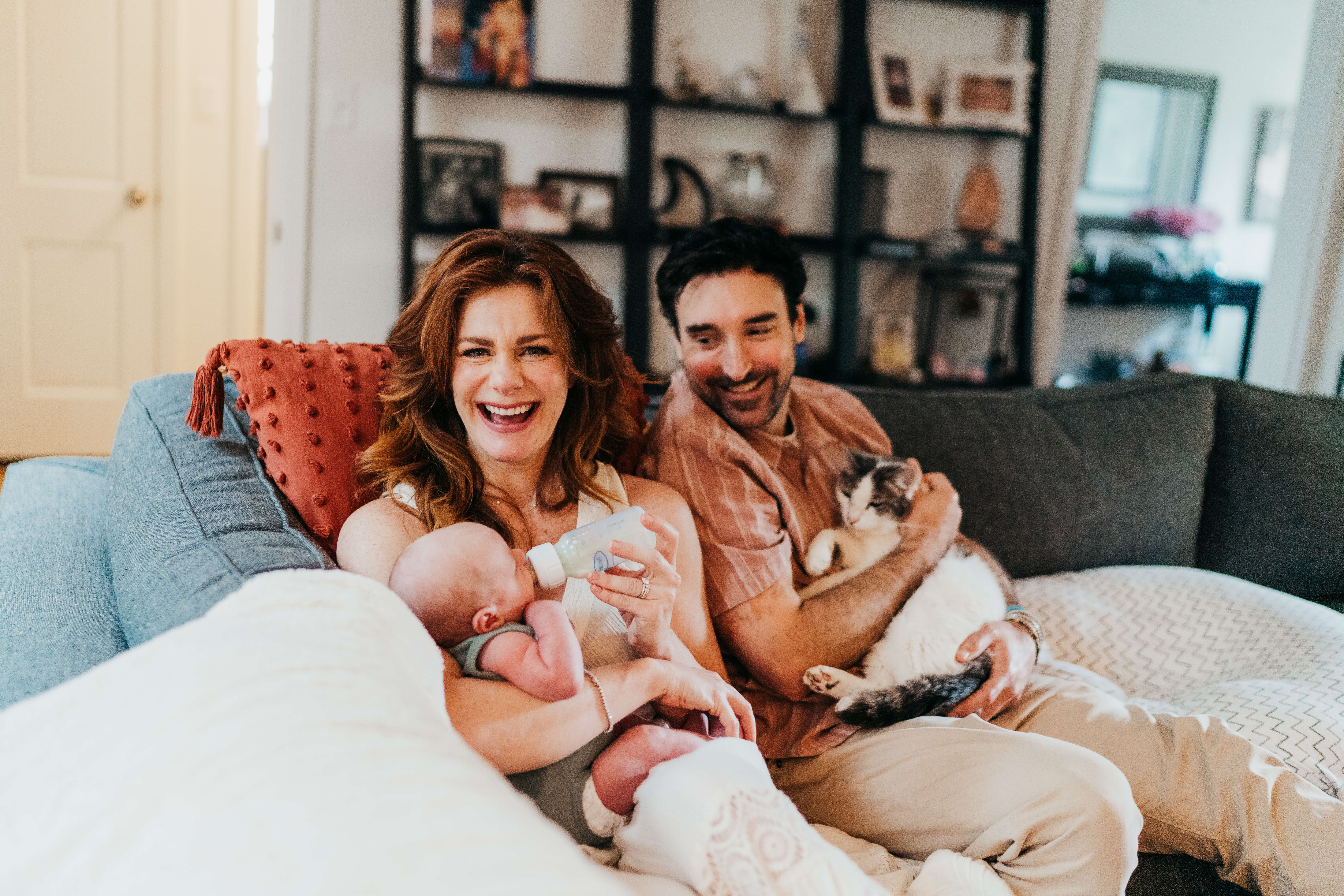 Happy new parents sit on a couch as dad cuddles a cat and mom feeds their newborn a bottle thanks to lactation consultants on mercer island