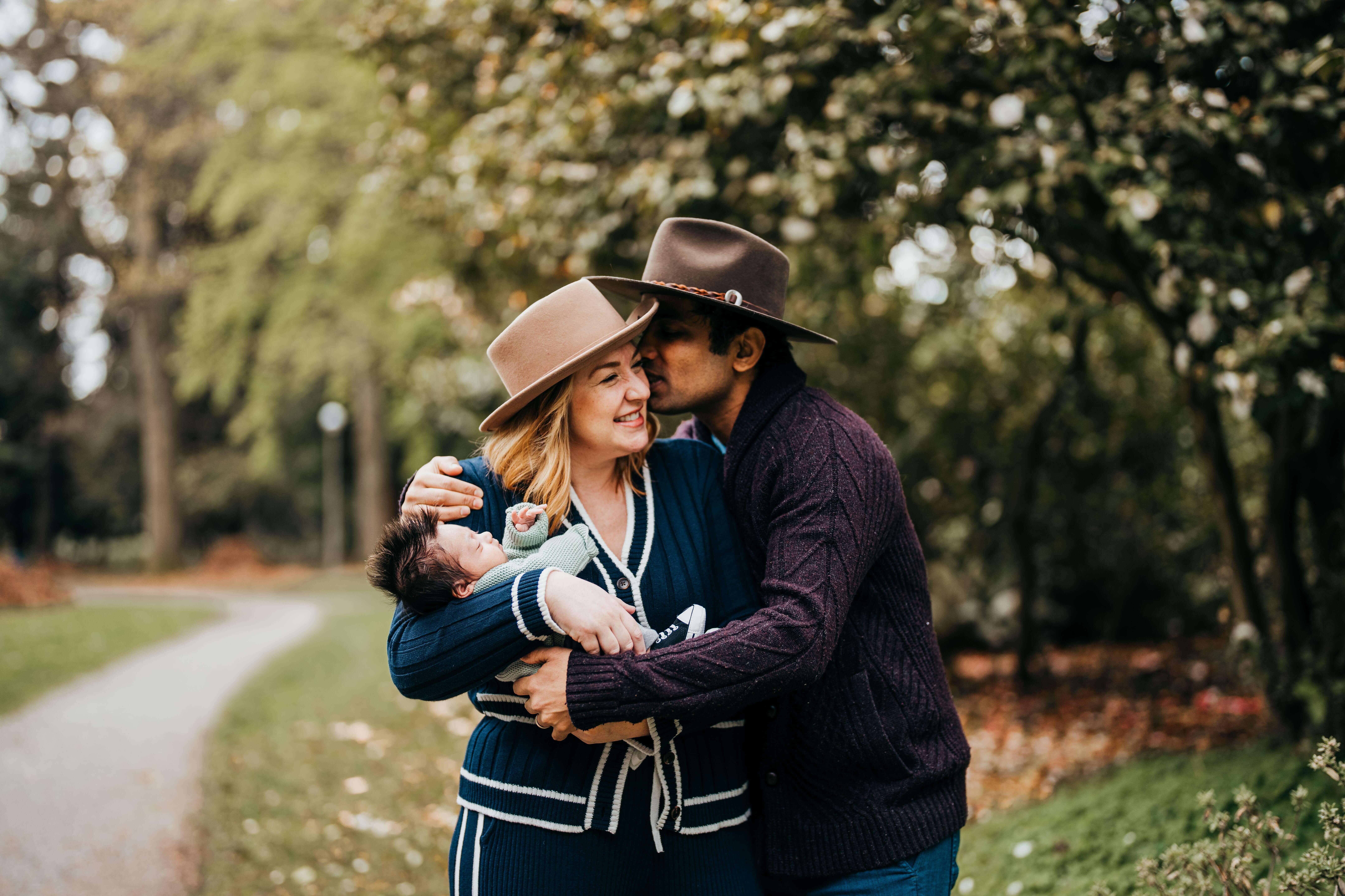 A dad in a purple sweater kisses the cheek of his happy wife as she holds their newborn in her arms in a blue sweater.