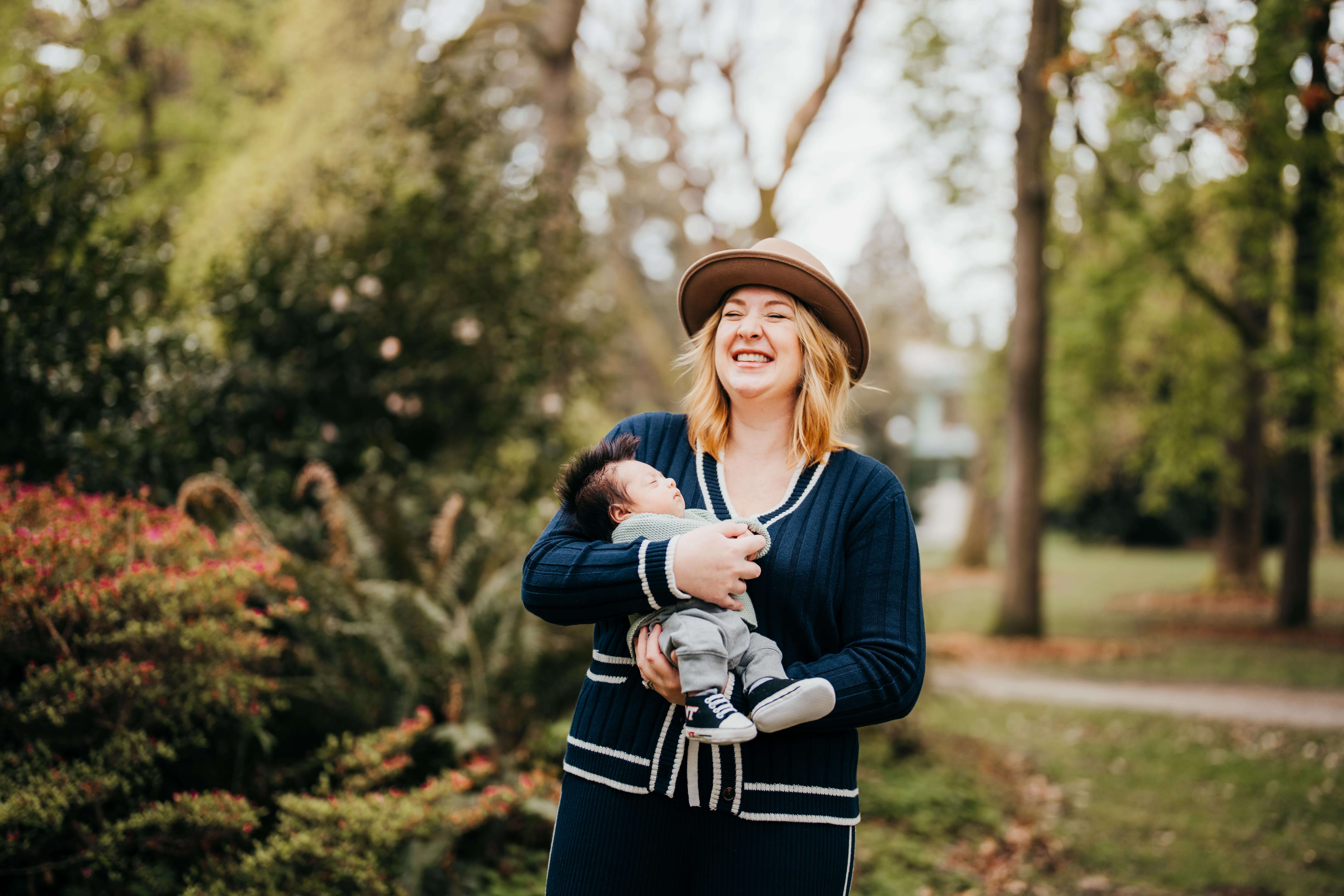 A giggling new mom in a blue sweater and tan hat holds her newborn in a onesie and shoes after visiting Chrysanthemum seattle