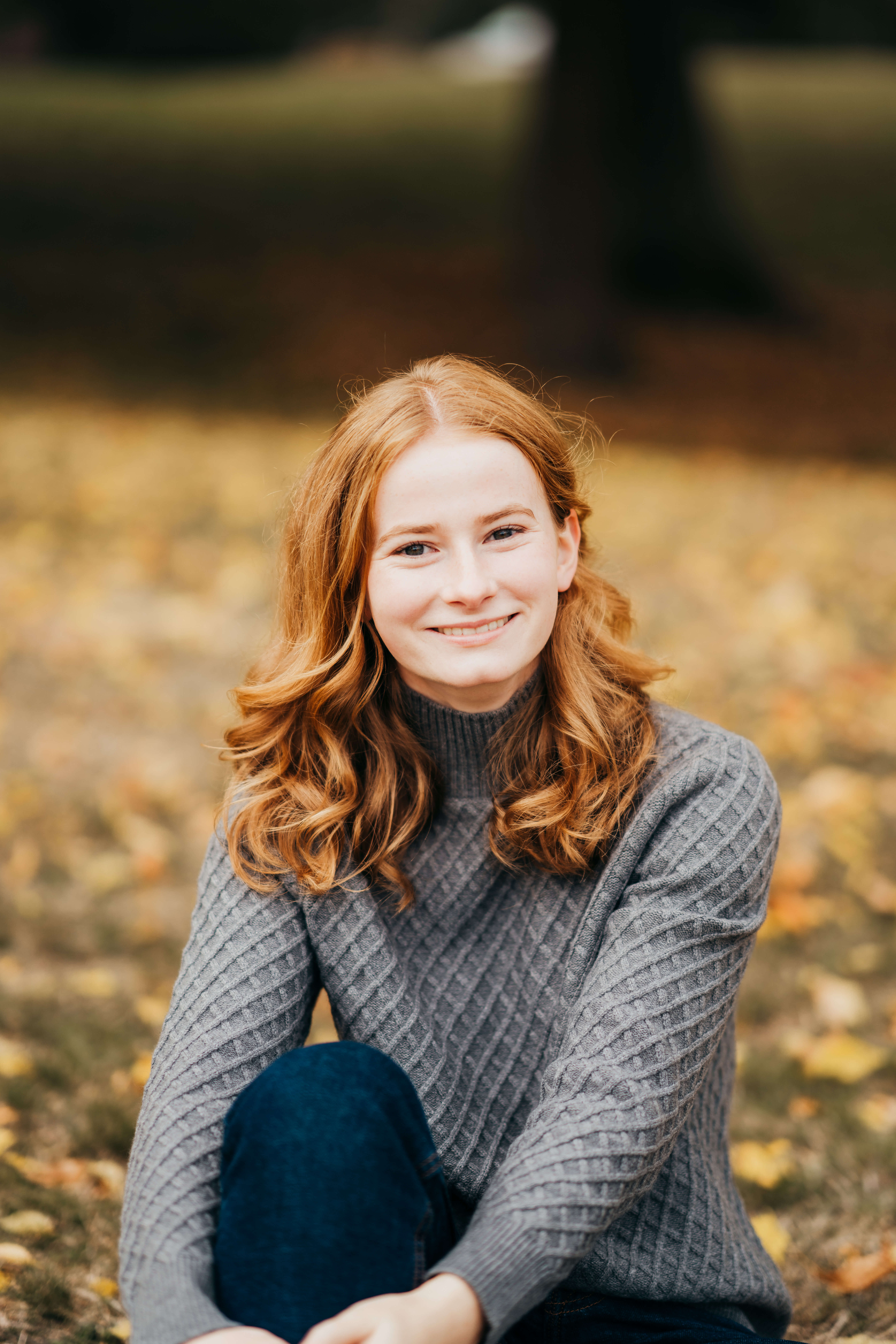 A happy red hair woman sits in a park lawn in a grey sweater