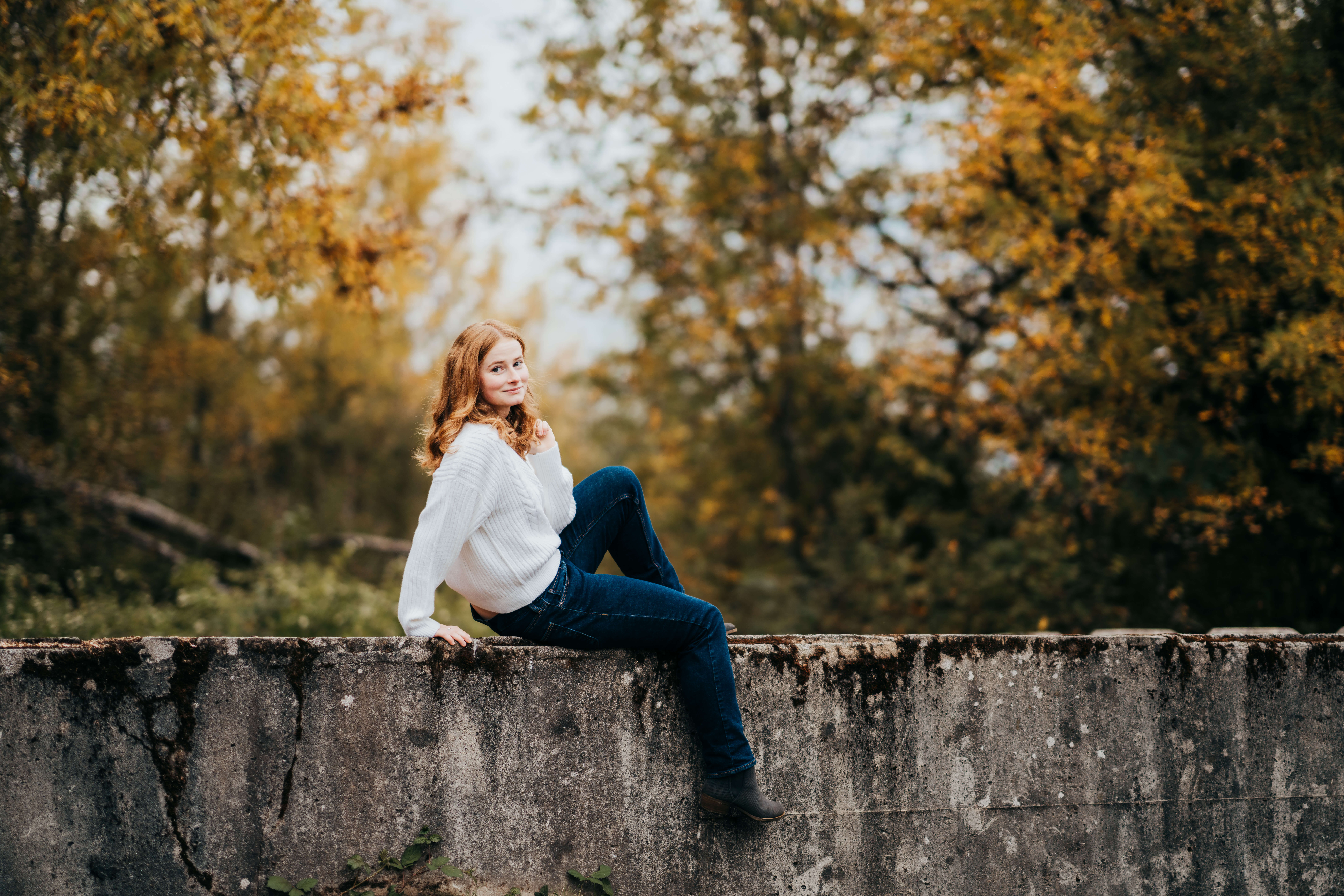 A happy woman in a white sweater and jeans sits on a cement wall in a forest after finding High School Volunteer Opportunities in Seattle