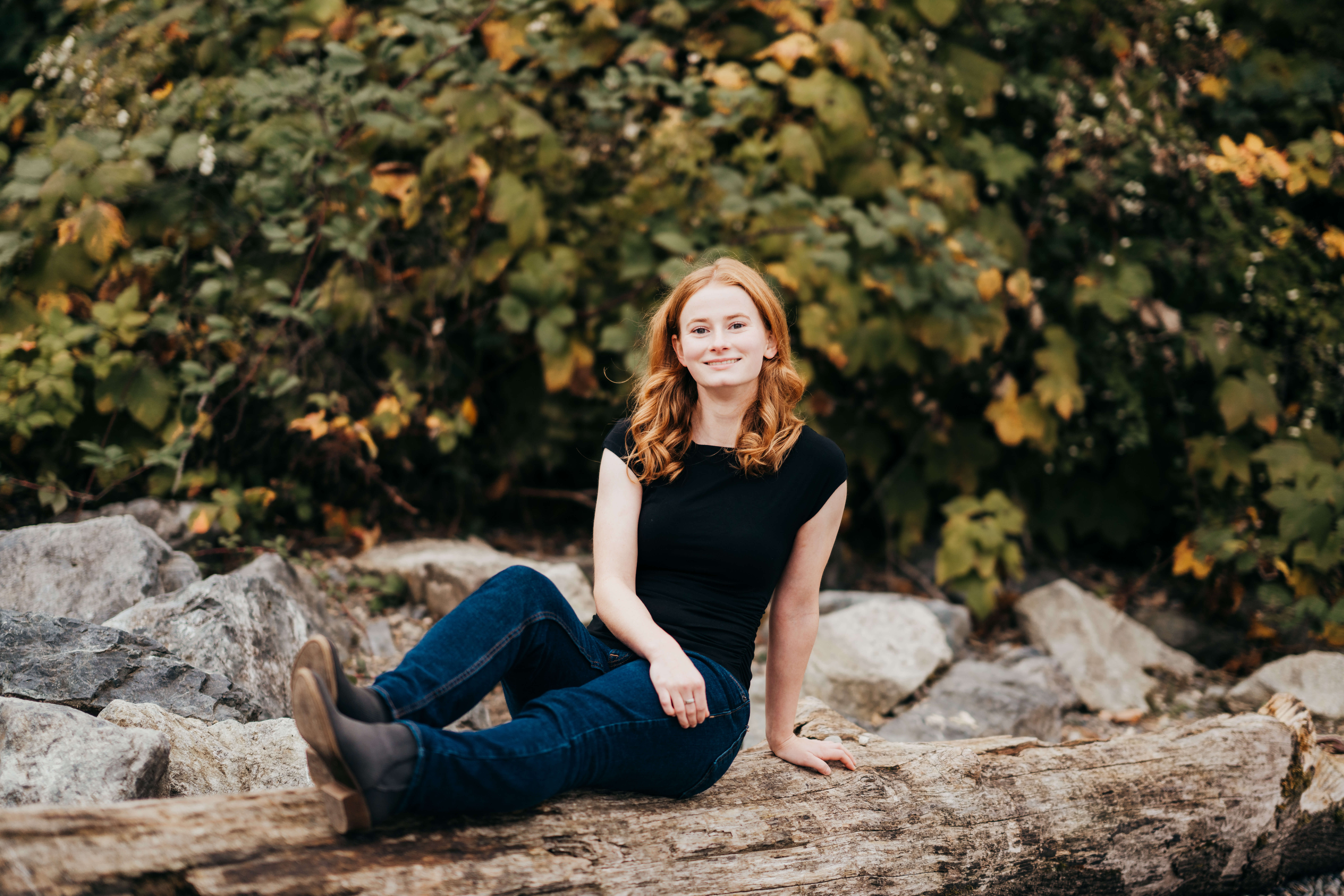 A teenage girl sits on a fallen log in a black shirt and jeans while smiling after finding High School Volunteer Opportunities in Seattle