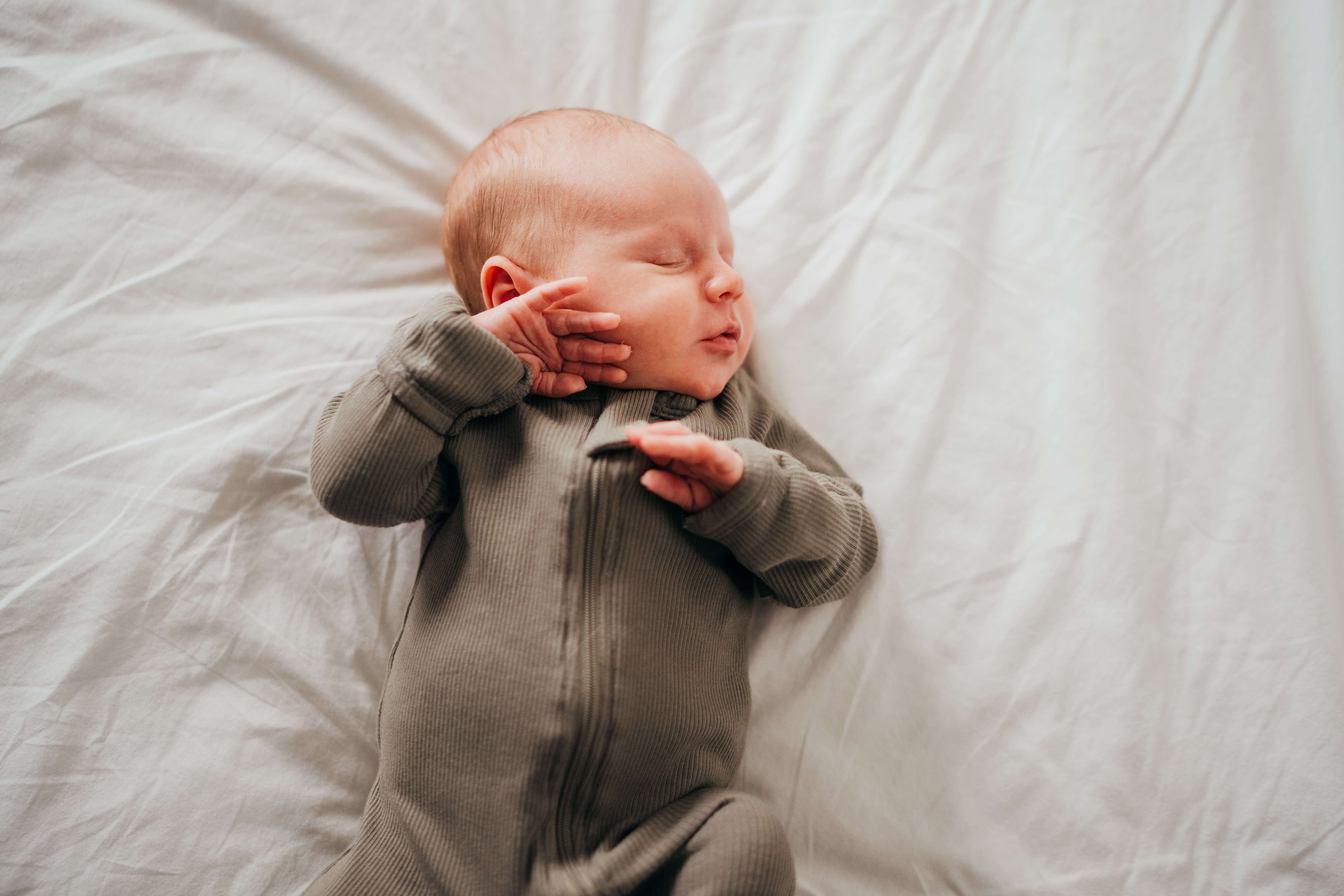 A sleeping newborn on a white bed in a green onesie with hands up