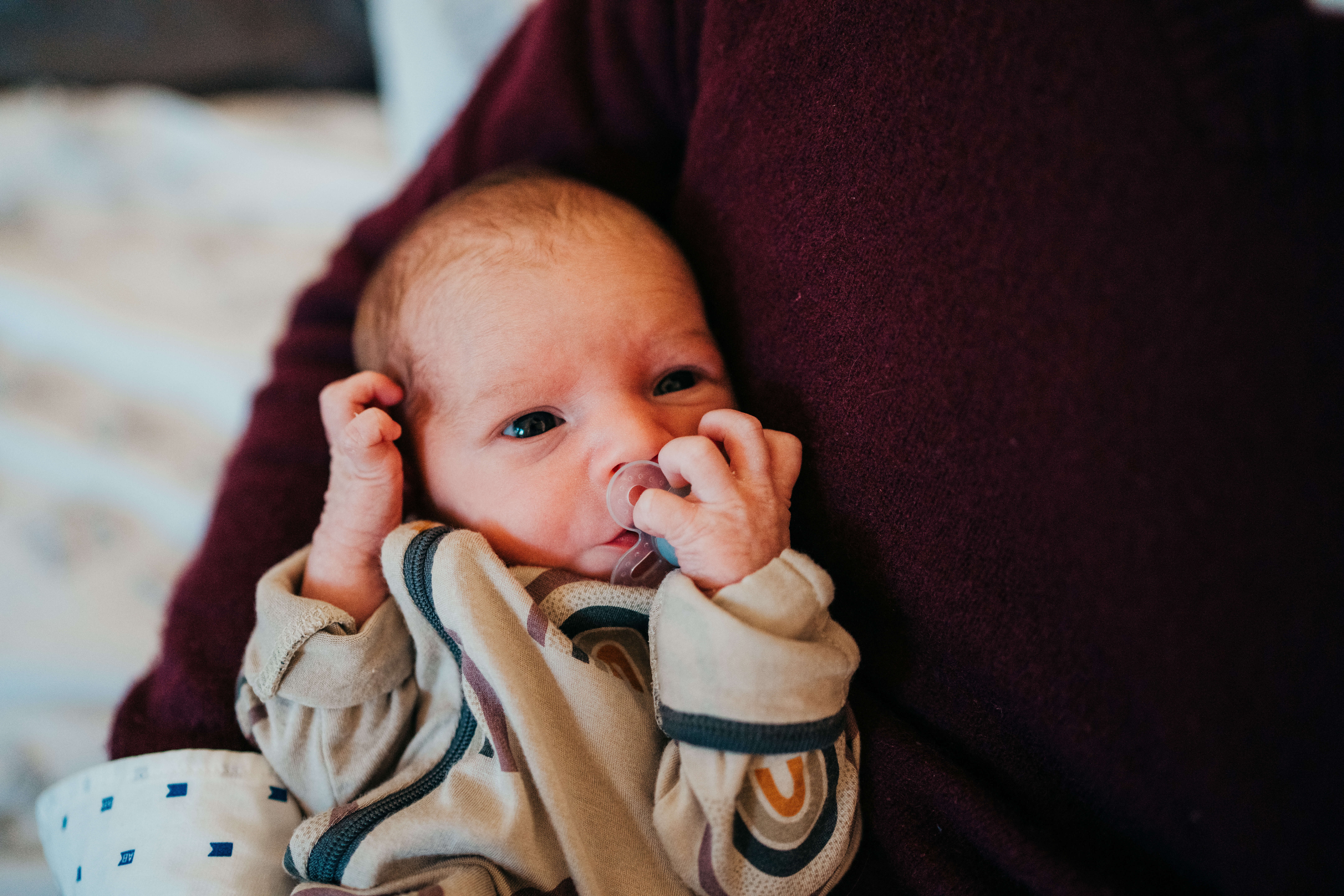 A newborn baby laying in parent's arms holding its binky with eyes open after visiting The Original Children's Shop