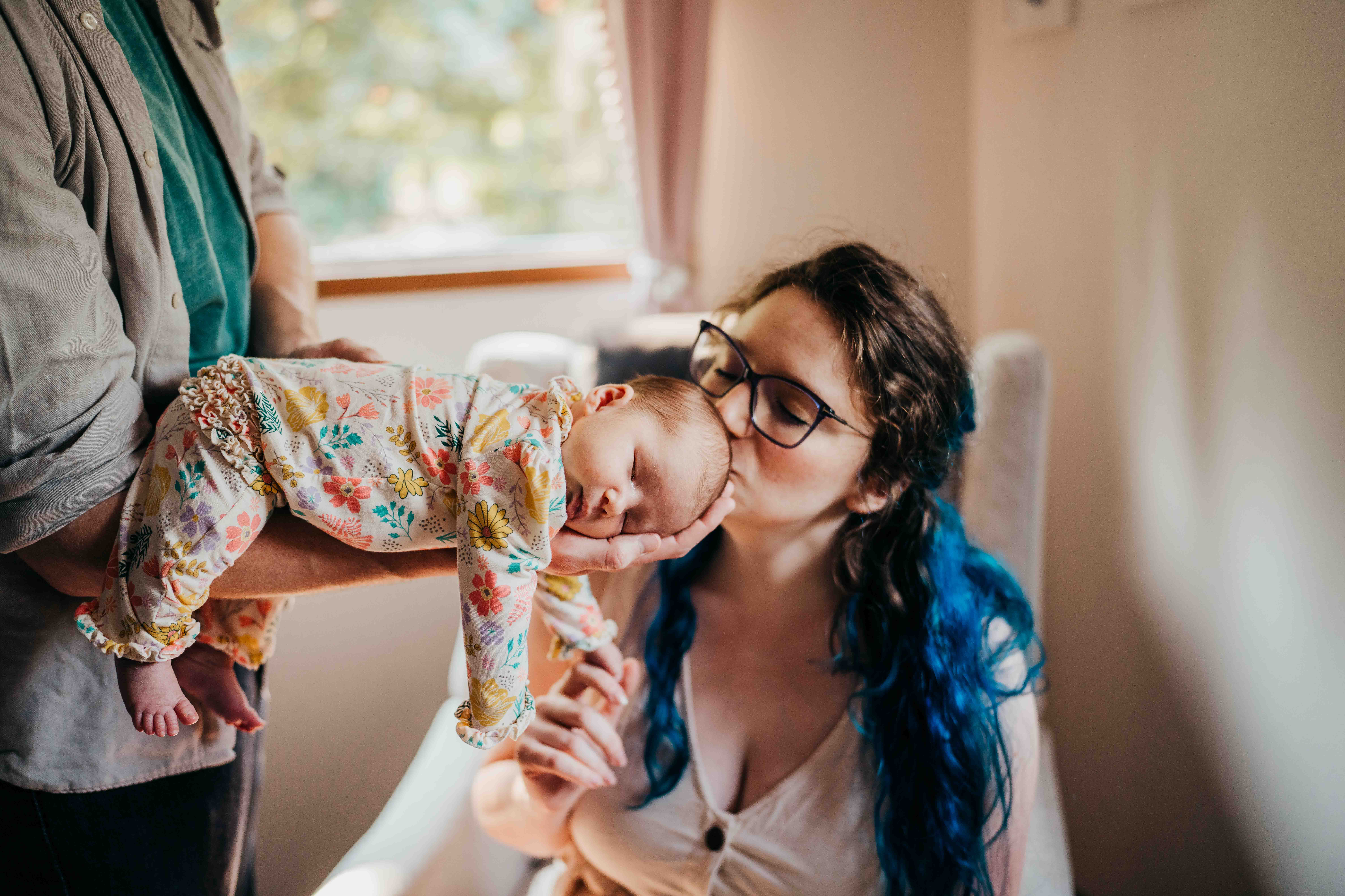 A newborn in a floral print onesie sleeps on dad's forearm as mom kisses her head while sitting in a nursing chair after exploring baby shower venues on mercer island