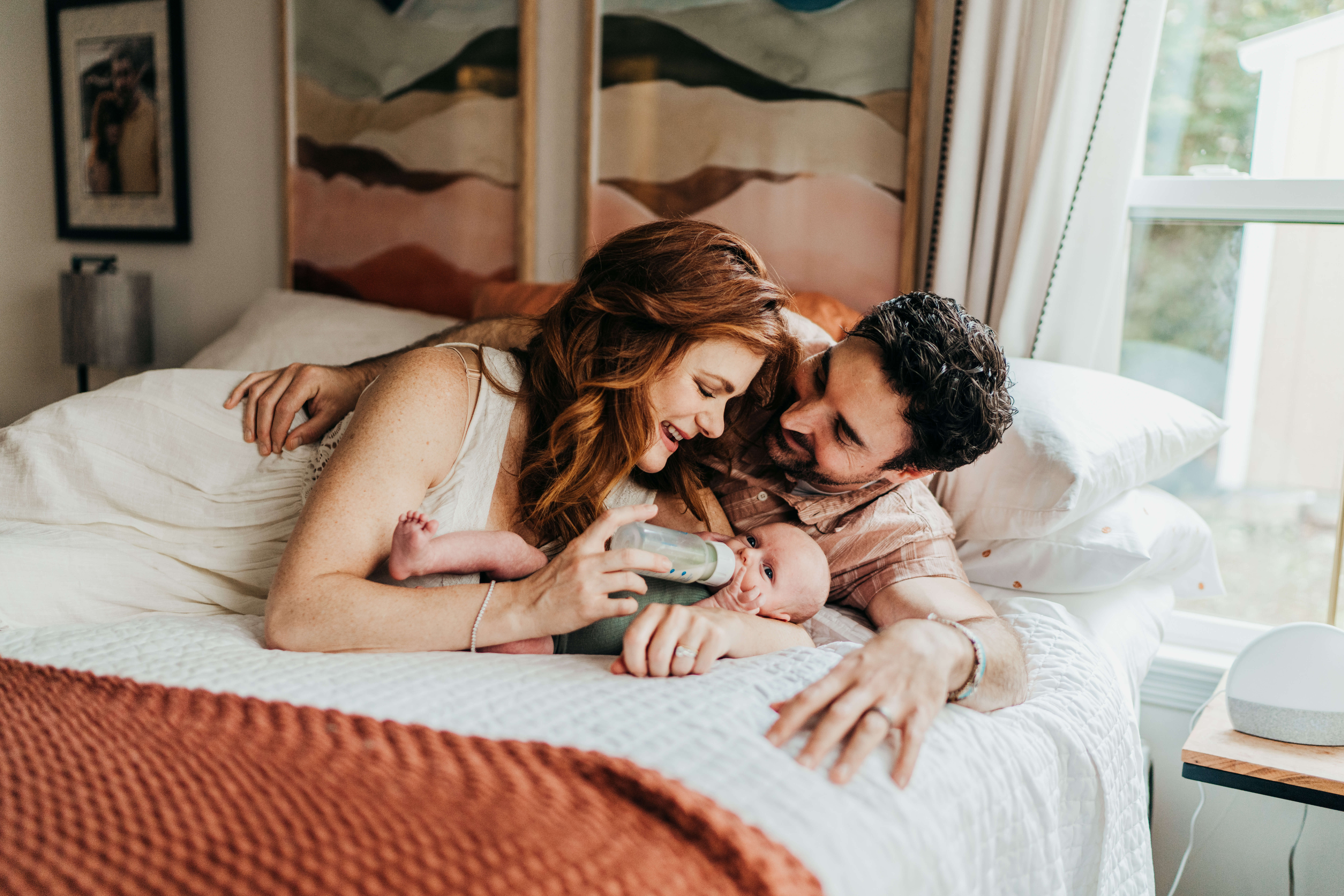 A mom and dad lay on a bed feeding their newborn a bottle while smiling and cuddling