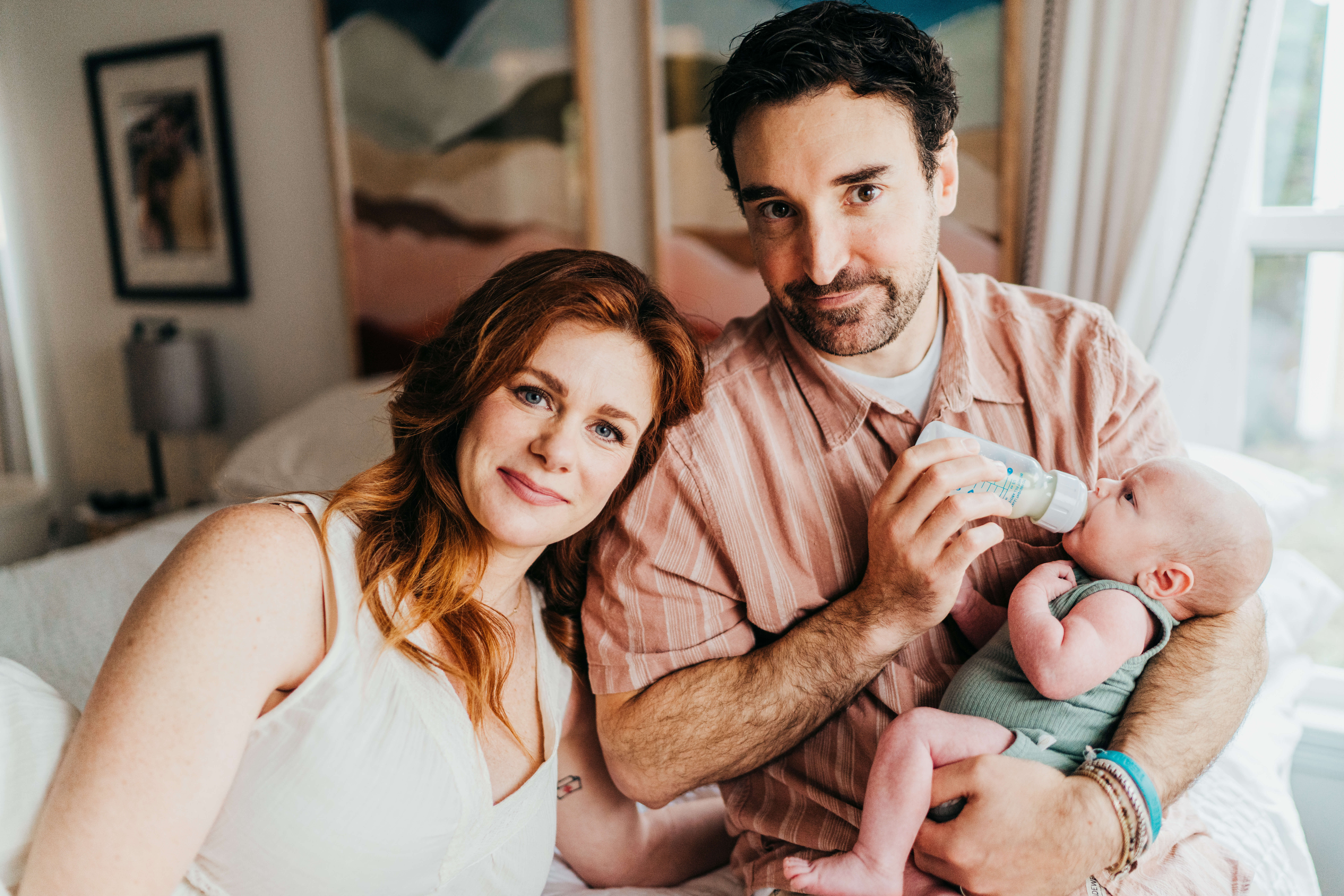Happy new parents snuggle while sitting on the edge of their bed feeding their infant from a bottle in a window thanks to ivf in seattle
