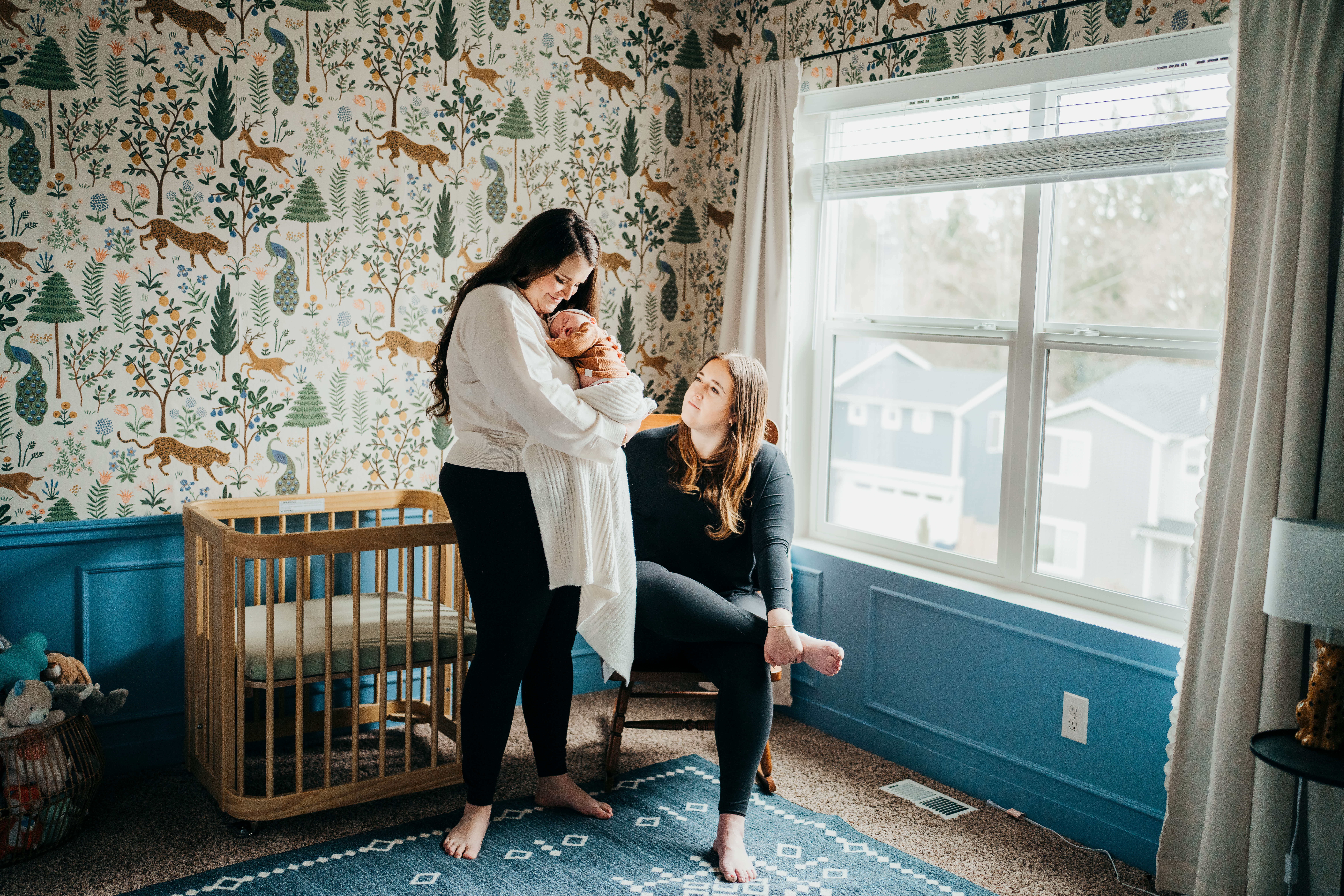 A happy new mom cradles her sleeping newborn girl on her chest while standing next to her wife sitting in a nursery in black