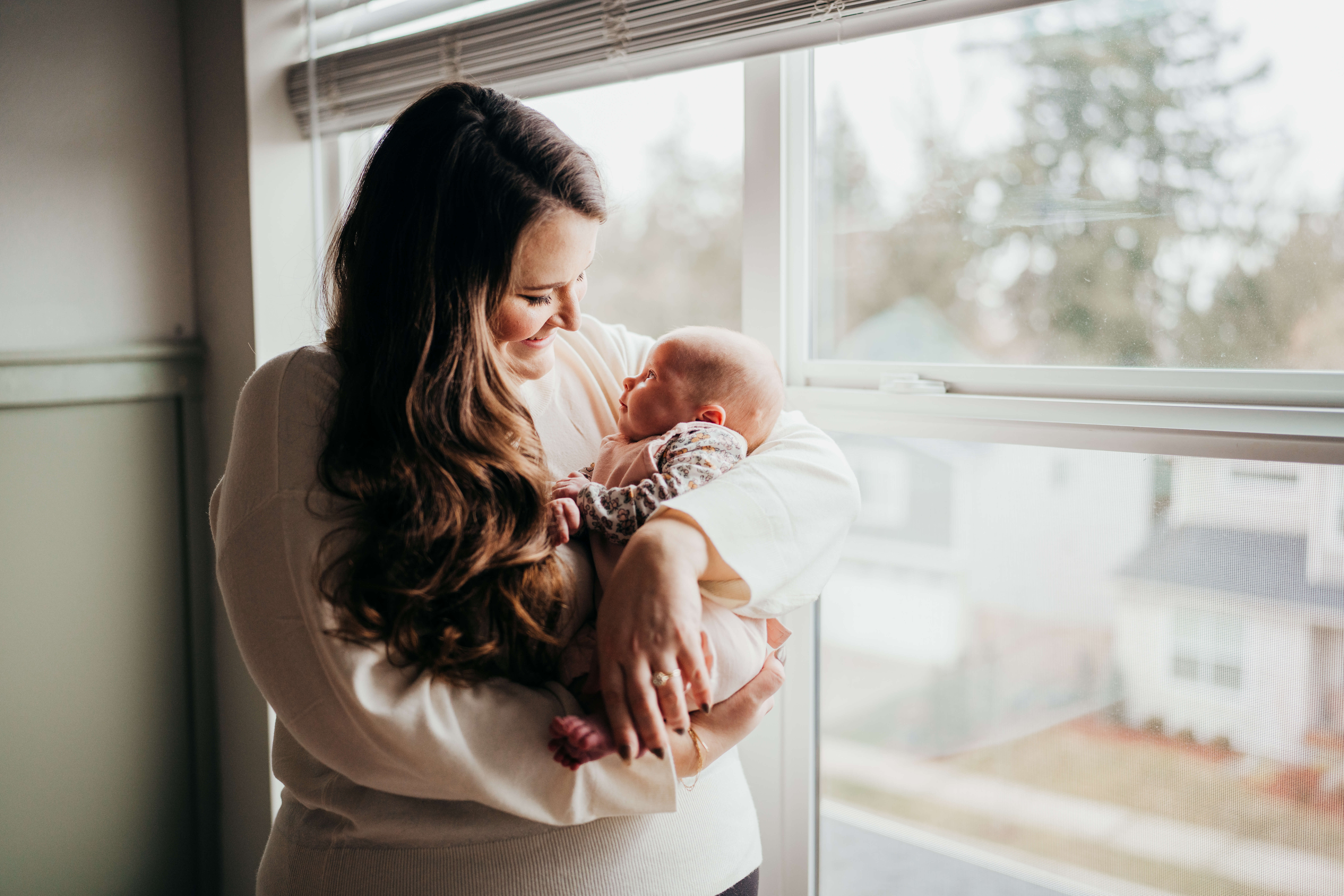 A smiling new mother in a white sweater gazes down at her newborn in her arms while standing in a window as baby looks up to her before finding pelvic floor therapy in seattle